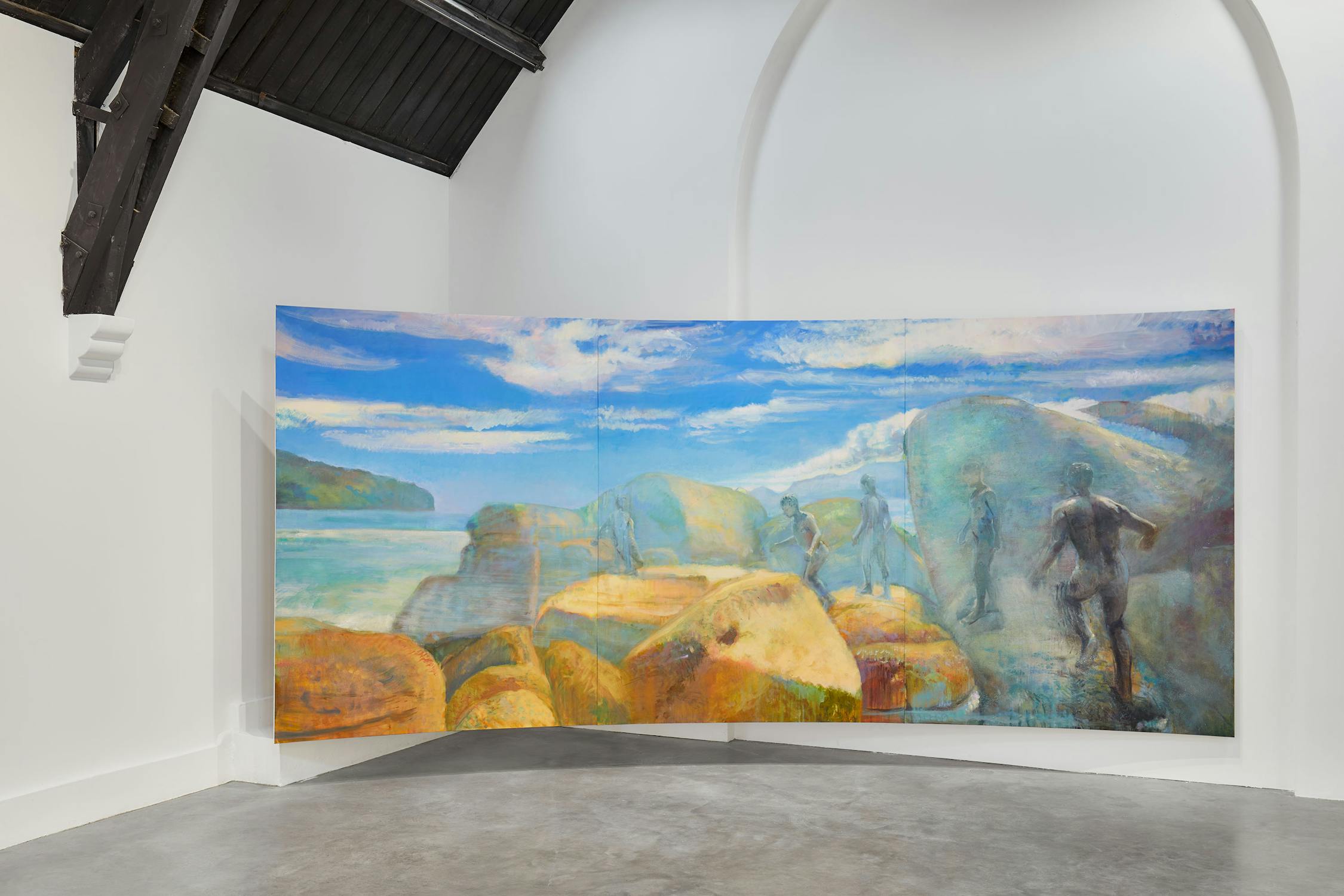 A large, colourful painting by Jake Grewal in a gallery depicts several abstract human figures moving on a rocky, beach-like landscape under a bright blue sky with clouds. The artwork is curved and displayed against white walls with a high ceiling.