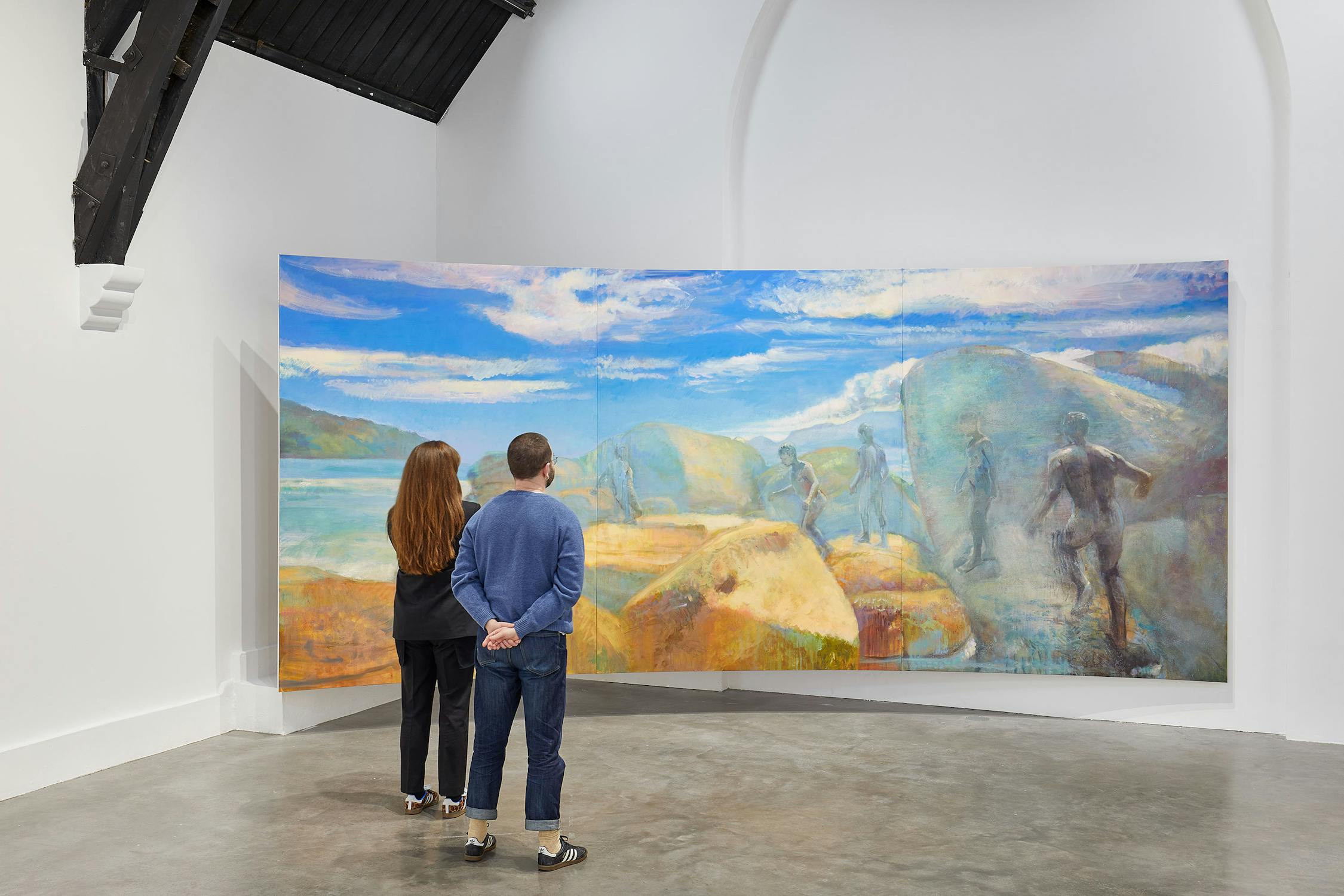 A gallery room with two paintings. On the left, a smaller painting with abstract colours. On the right, a large mural depicting figures running on rocks by a body of water, under a partly cloudy sky. The room has high, dark, vaulted ceilings.