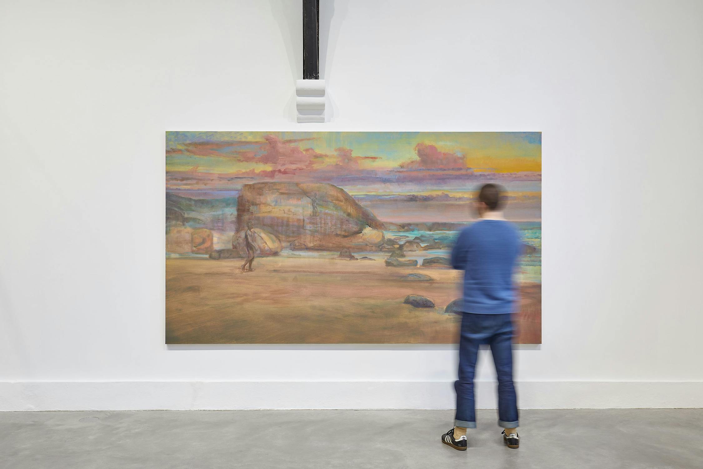 A person wearing a blue sweater and jeans stands in front of a large painting depicting a rocky beach scene with waves and a colourful sky displayed on a gallery wall.