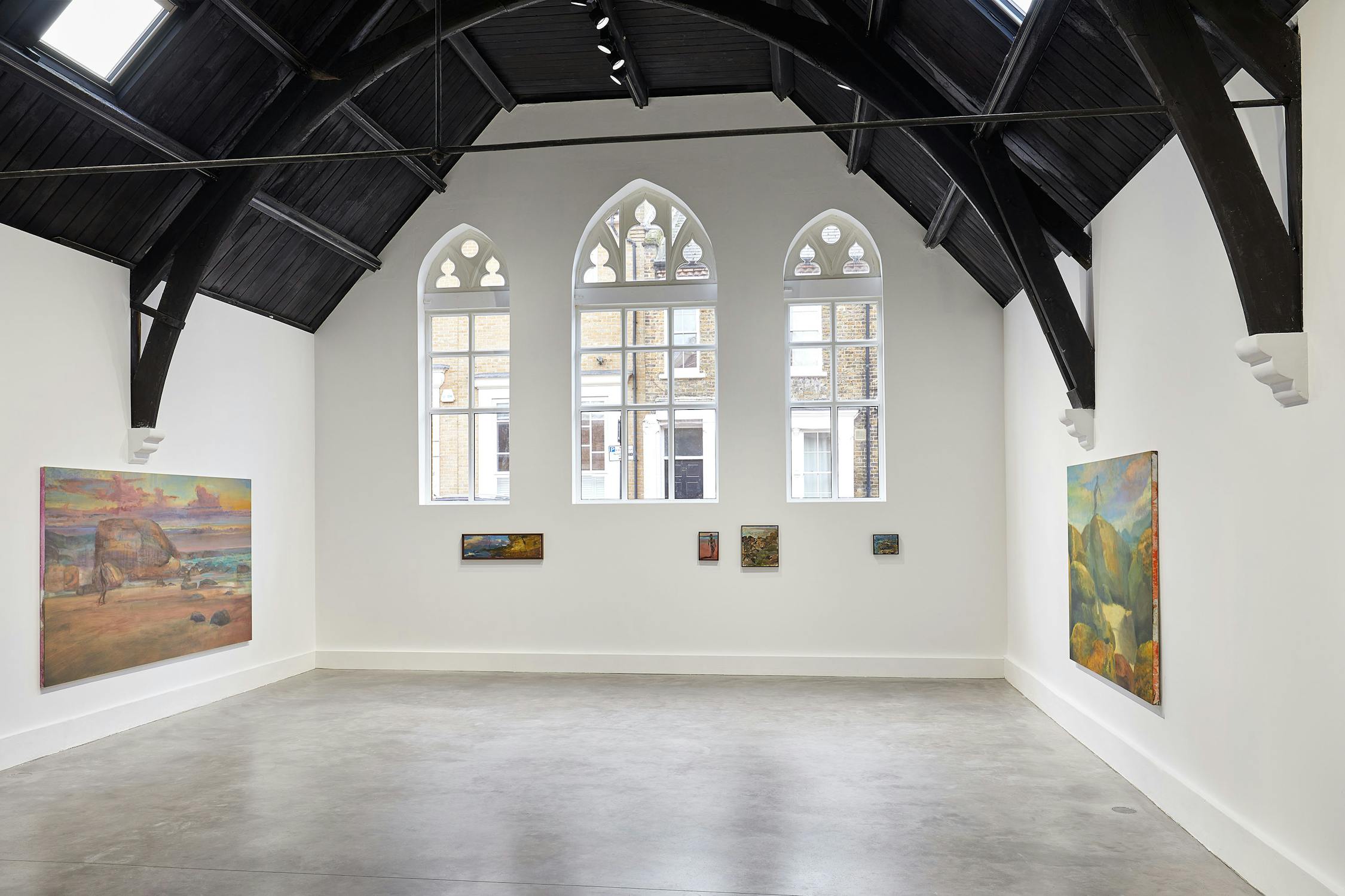 A spacious art gallery with a high, dark wood ceiling features several landscape paintings by Jake Grewal on white walls. Large arched windows let in natural light, highlighting the paintings. The floor is a polished grey concrete.