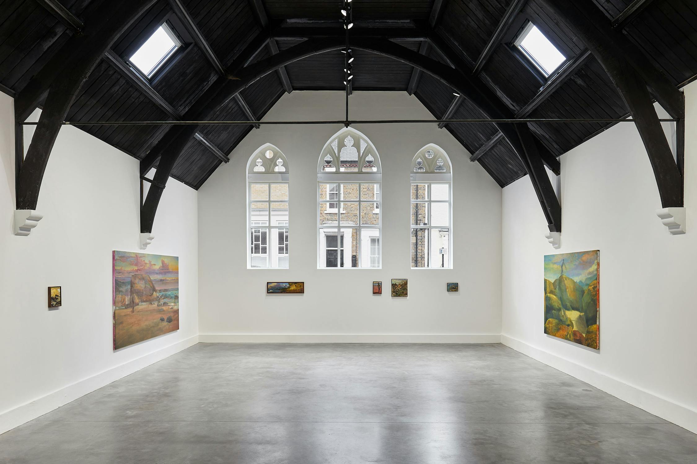 Spacious art gallery with high wooden ceiling and arched windows, featuring landscape paintings by Jake Grewal on white walls. Natural light streams in, illuminating the polished concrete floor.