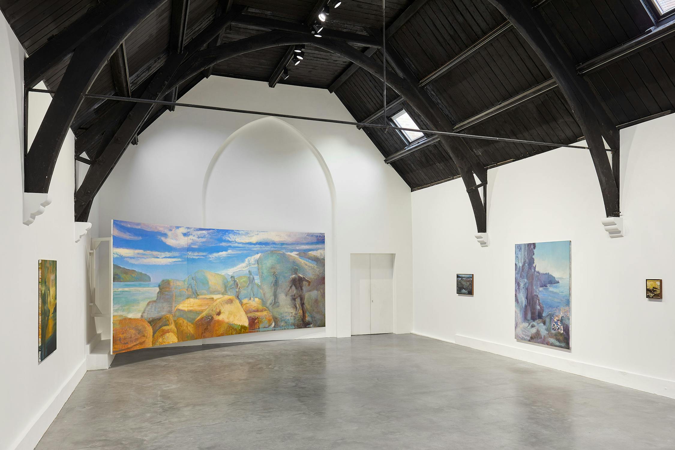 A spacious art gallery with a high, dark wooden ceiling features several landscape paintings by Jake Grewal on white walls. The large central painting depicts a vibrant coastal scene with rocks and blue skies. The gallery floor is polished concrete.