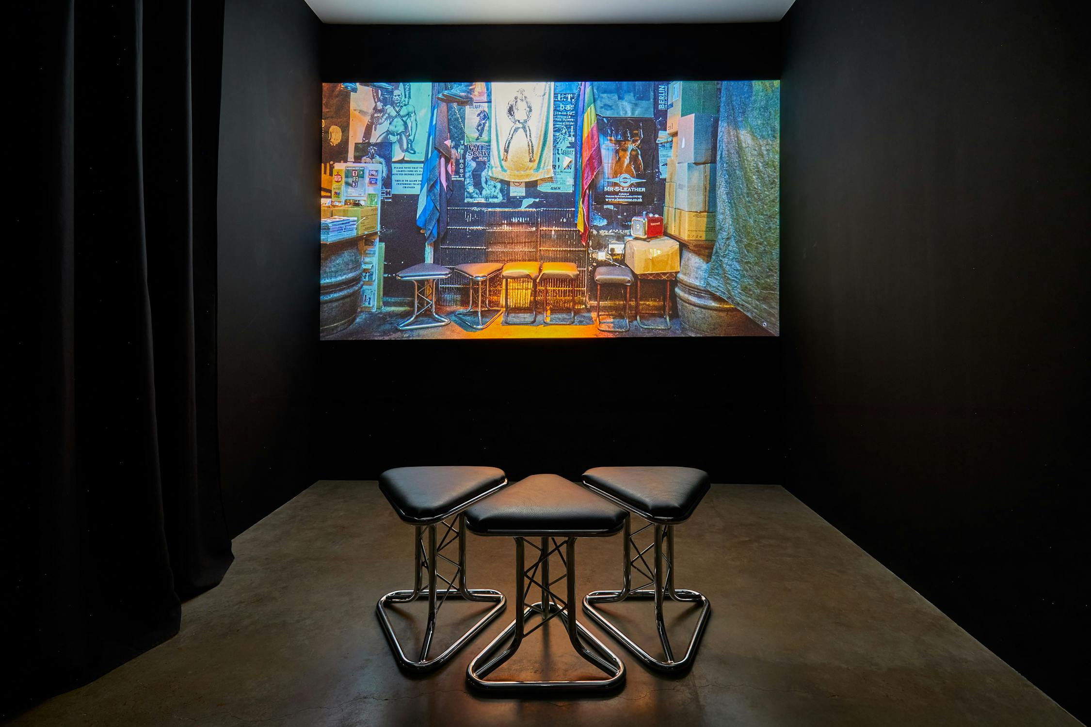 Three triangular stools face a large screen displaying a dimly lit room with empty stools and posters. The setup suggests an immersive exhibition or presentation in a dark, minimalist space.