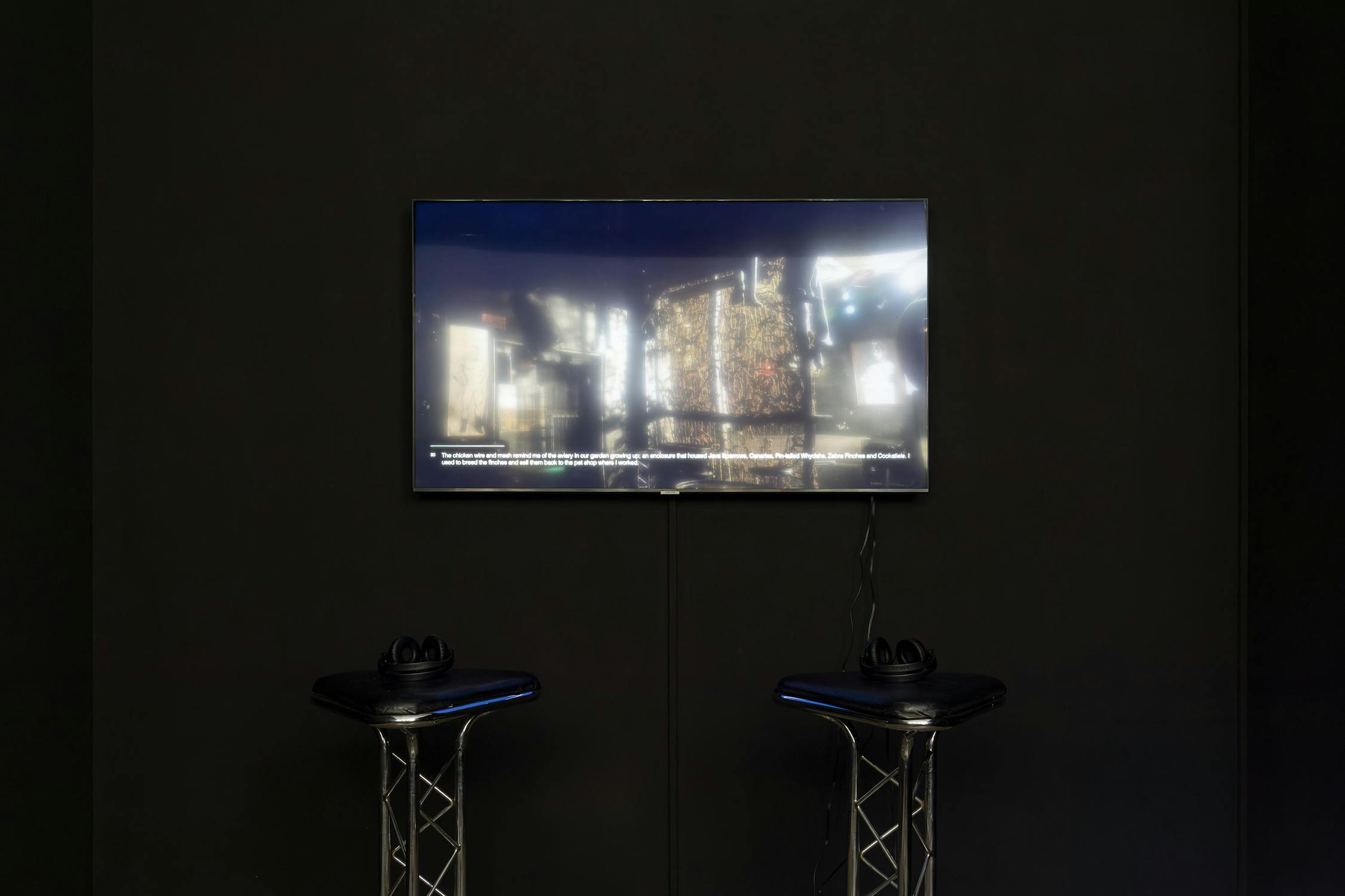 A television mounted on a dark wall displays a blurred image with text at the bottom. Two black stools with headphones rest on metal stands in front of the TV.
