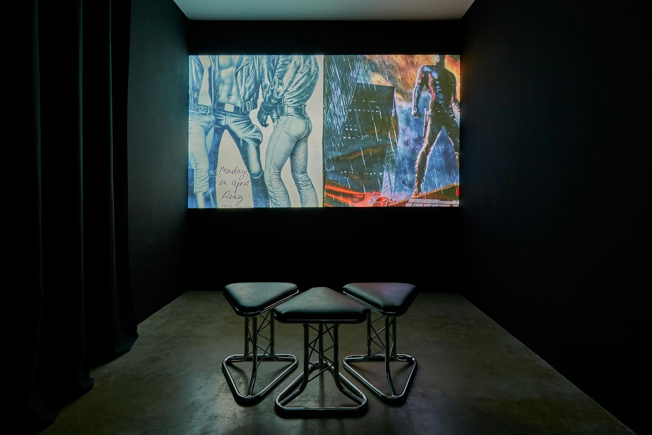 Three black stools face a large, illuminated screen displaying artistic images of people in jeans and abstract backgrounds. The room is dark, with textured flooring and black walls, creating a moody atmosphere.