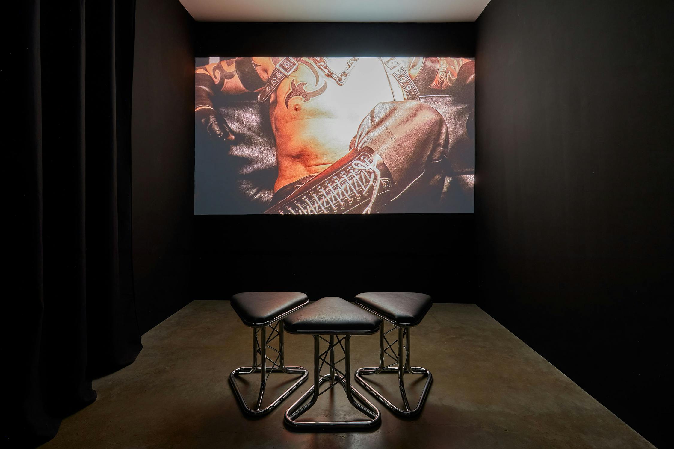 Three black stools are arranged in front of a large screen displaying a close-up image of a person in leather attire with visible tattoos and chains. The room has dark walls and a concrete floor.