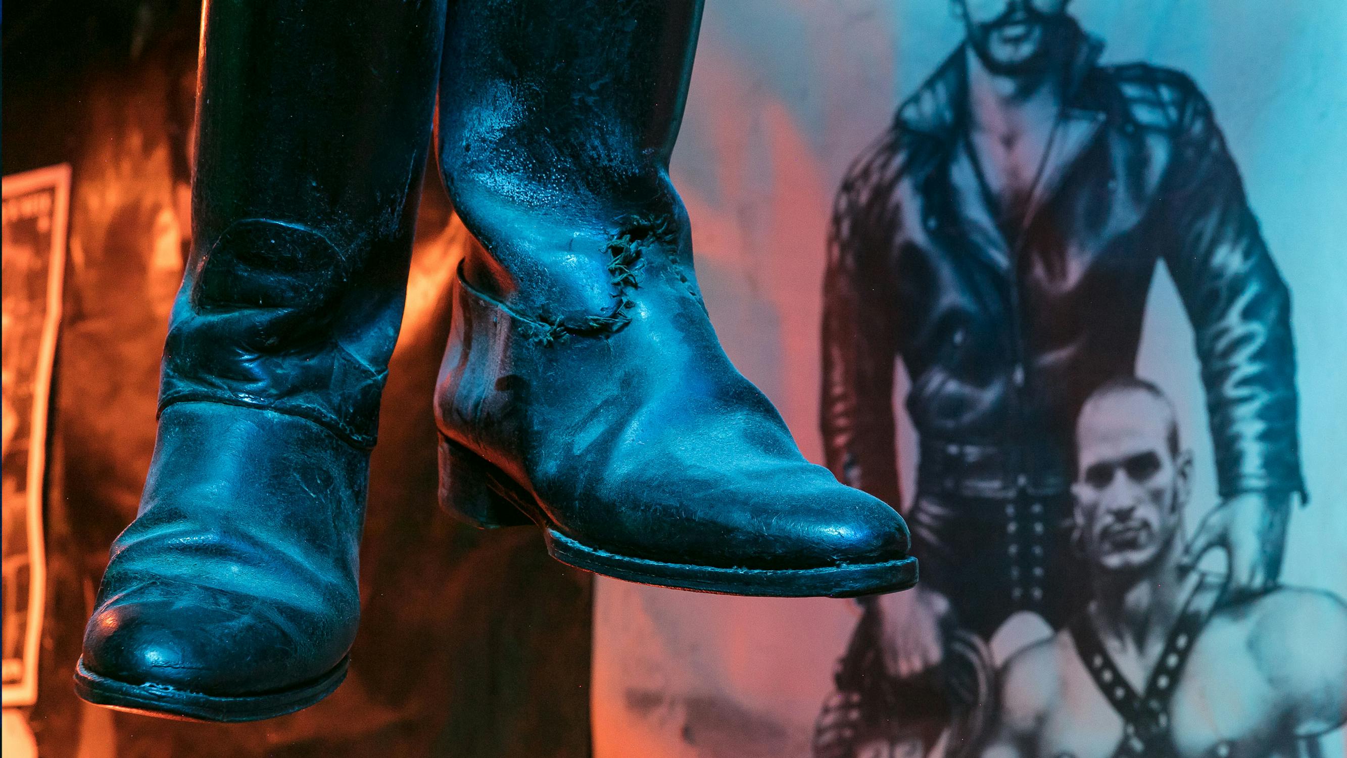 Worn black boots hanging with a dimly lit background, featuring a poster on the wall depicting men in leather attire, blending with the blue and orange lighting.