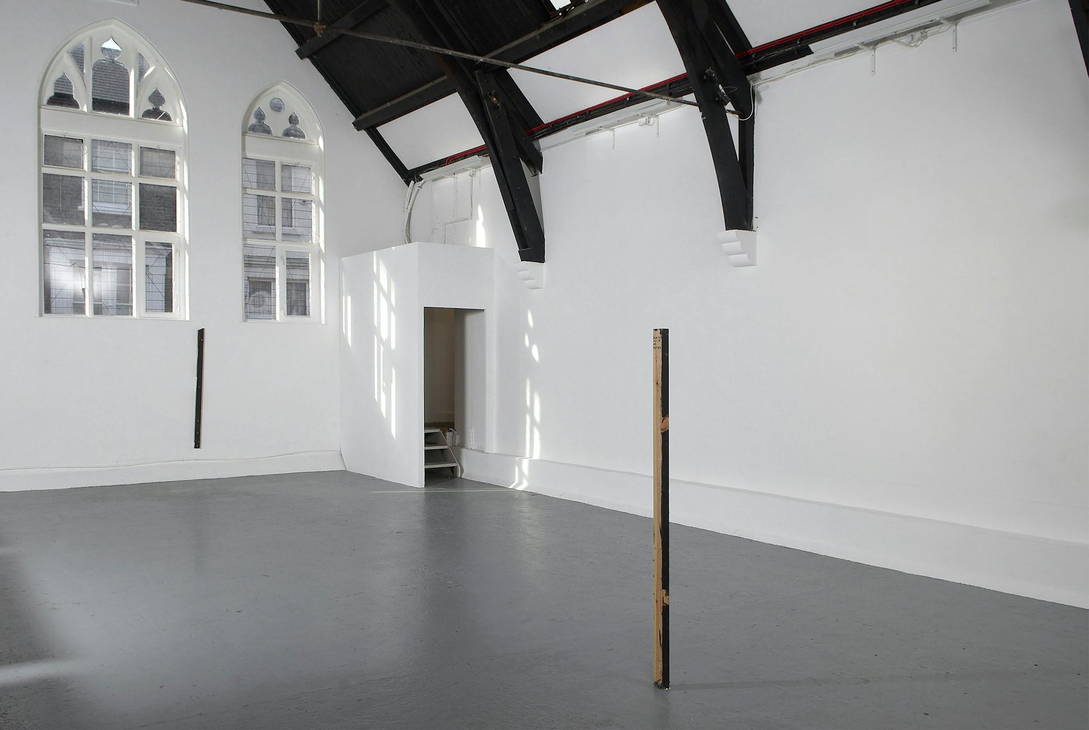 A minimalist gallery with arched windows casting light onto white walls and a grey floor. There are three vertical wooden posts installed in the space, and an open doorway leading to another area. Black beams line the ceiling.