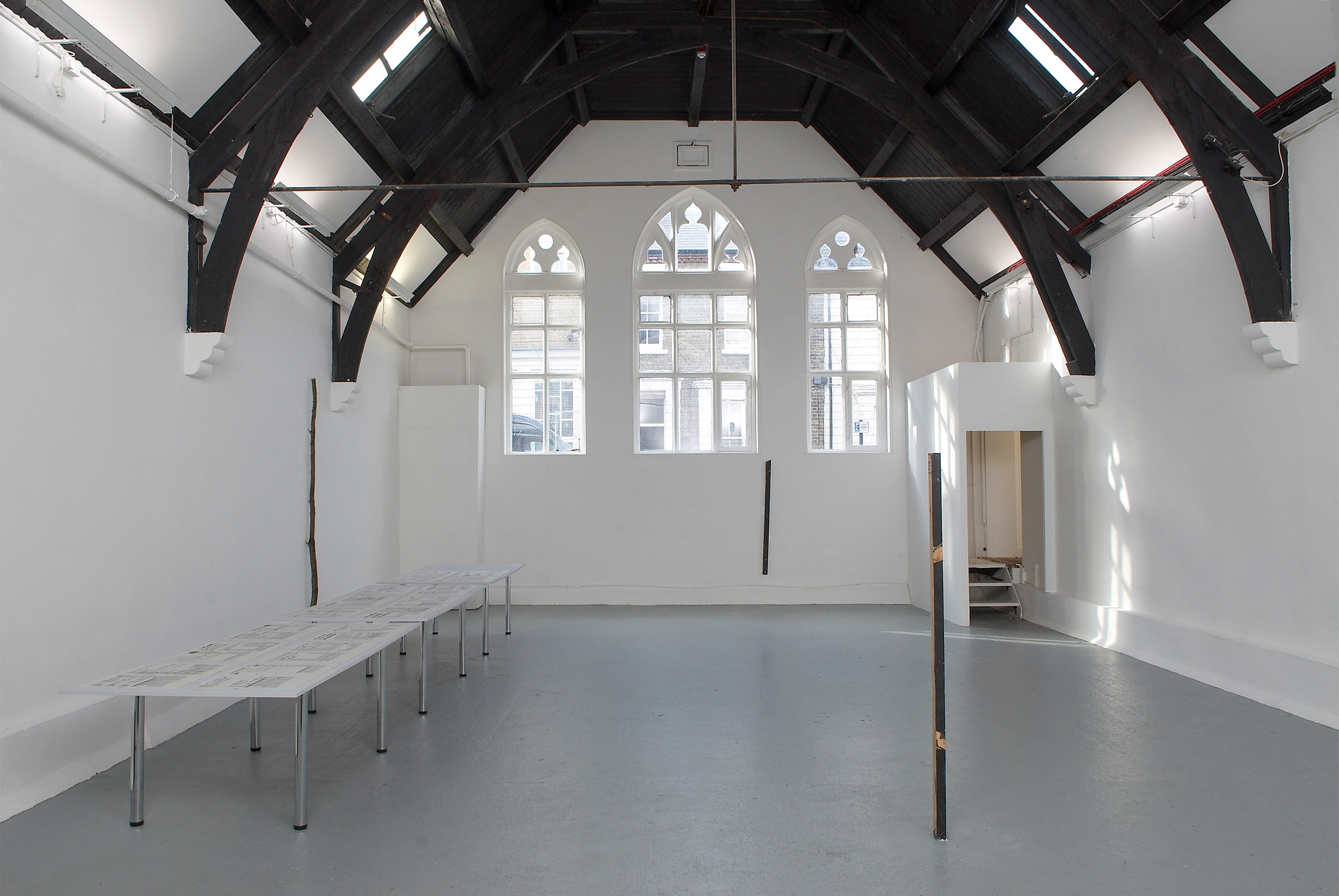 A spacious gallery with high, dark wooden beams and arched windows allowing natural light to enter. The walls and floor are white. A long table stands on the left, and there is a small doorway on the right.
