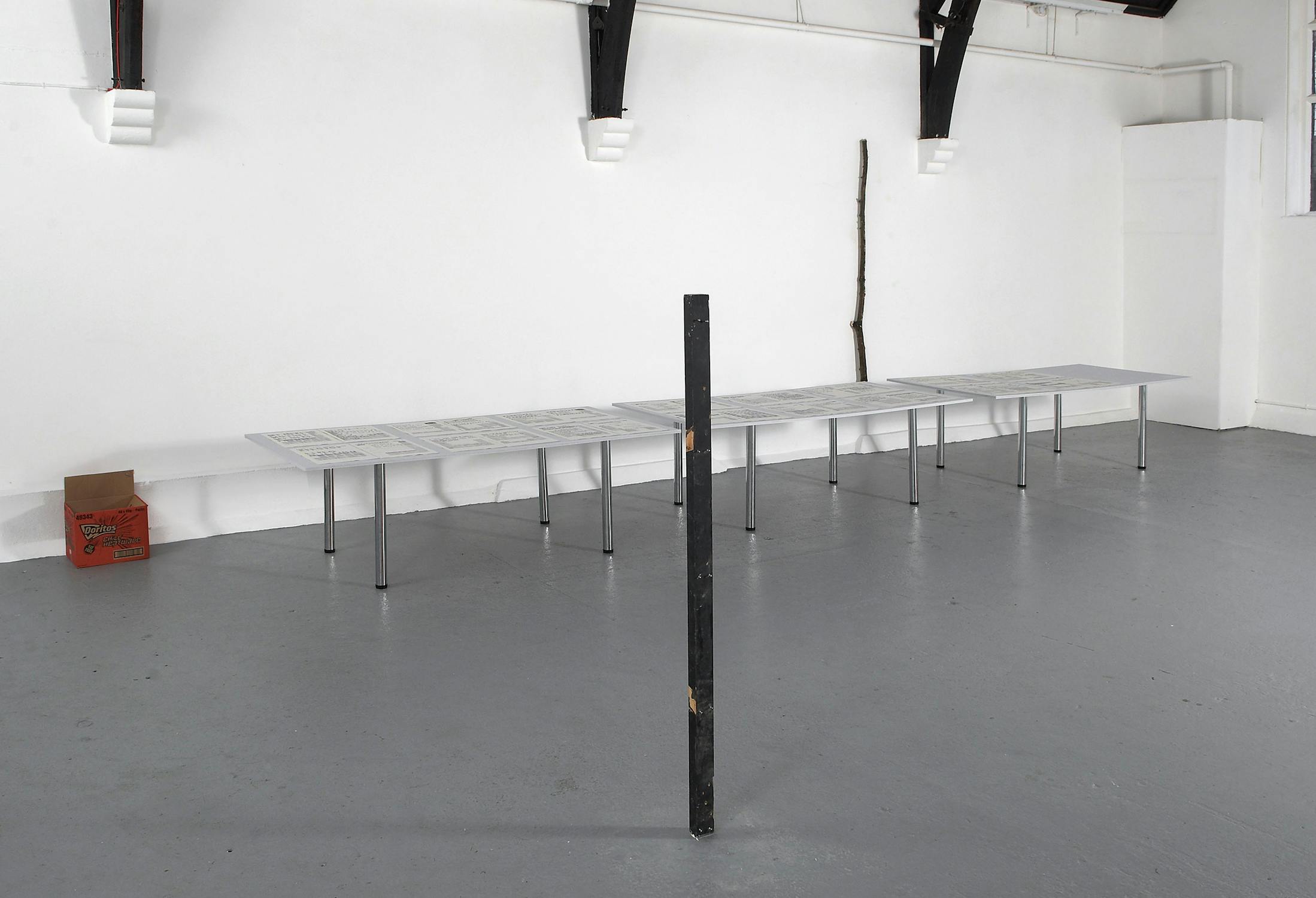 A minimalist art gallery display features a long table with papers, a slender vertical black pole in the foreground, and a solitary stick leaning against a wall. A cardboard box sits in the corner of the grey, open space.
