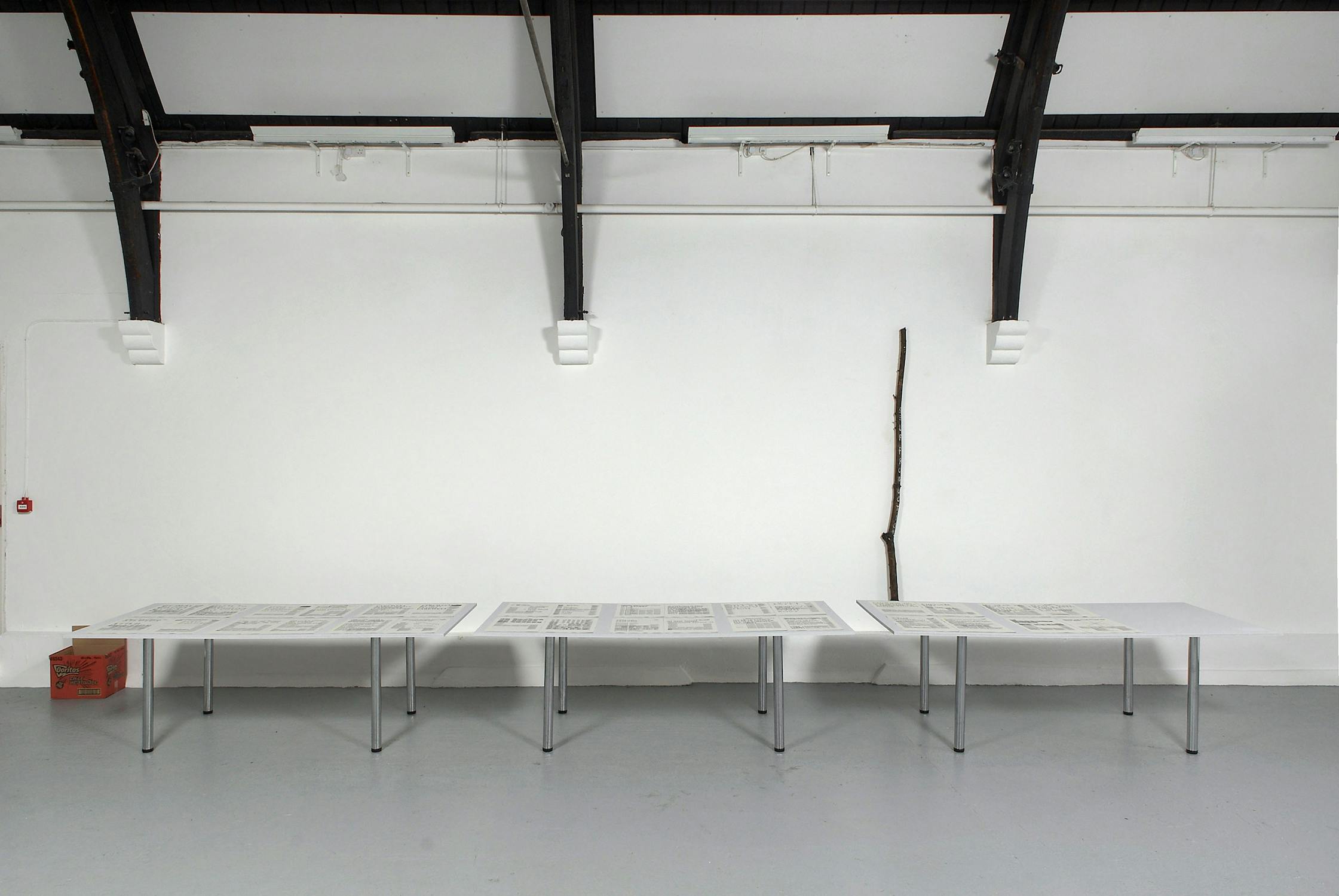 A minimalist gallery space with a high ceiling features three tables displaying newspapers. A single vertical branch leans against the white wall, and an orange box is placed on the floor to the left.