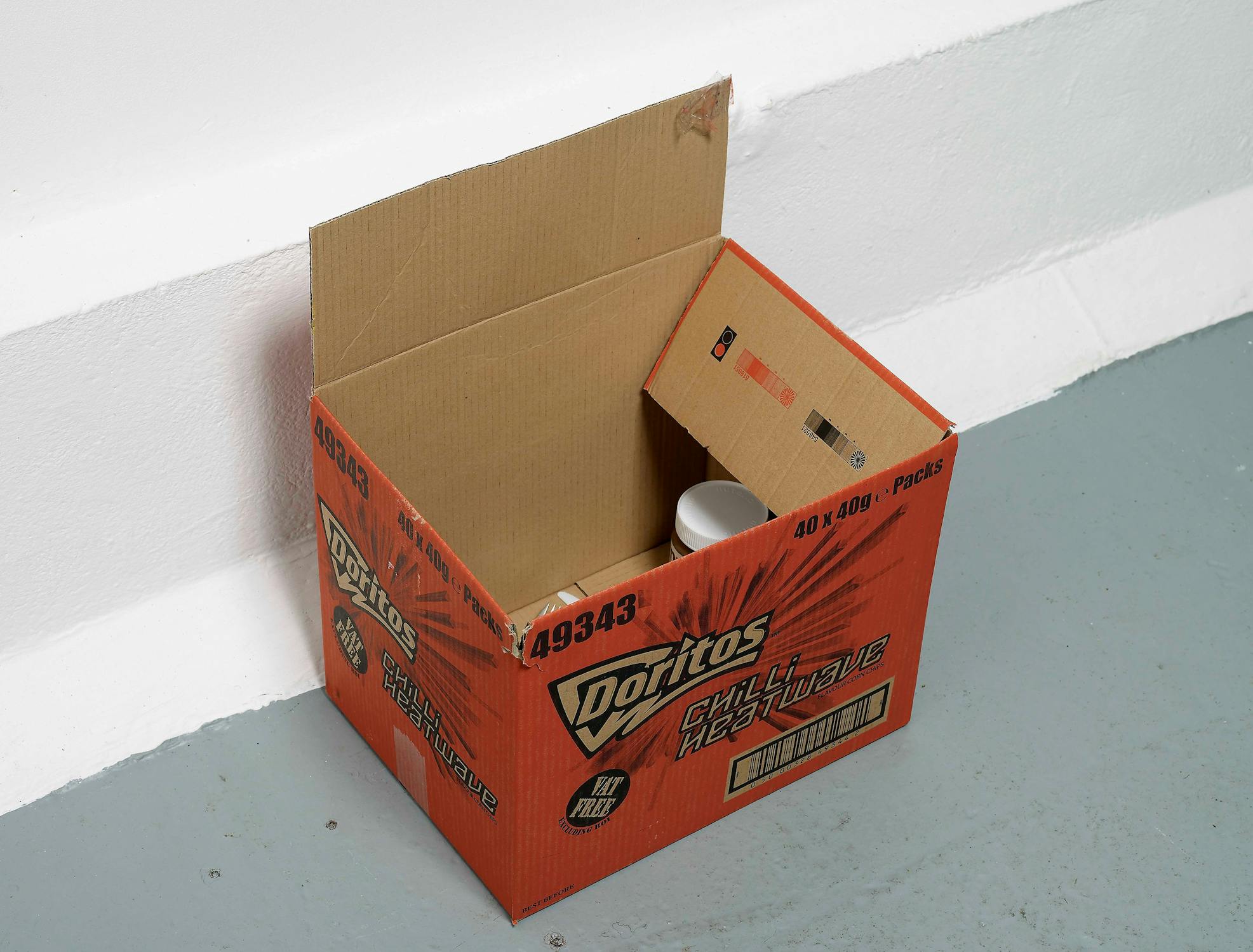 An open red Doritos box labeled "Chili Heatwave" is on the floor against a white wall. Inside the box, there is a white mug. The box is partially folded, revealing its interior flaps.