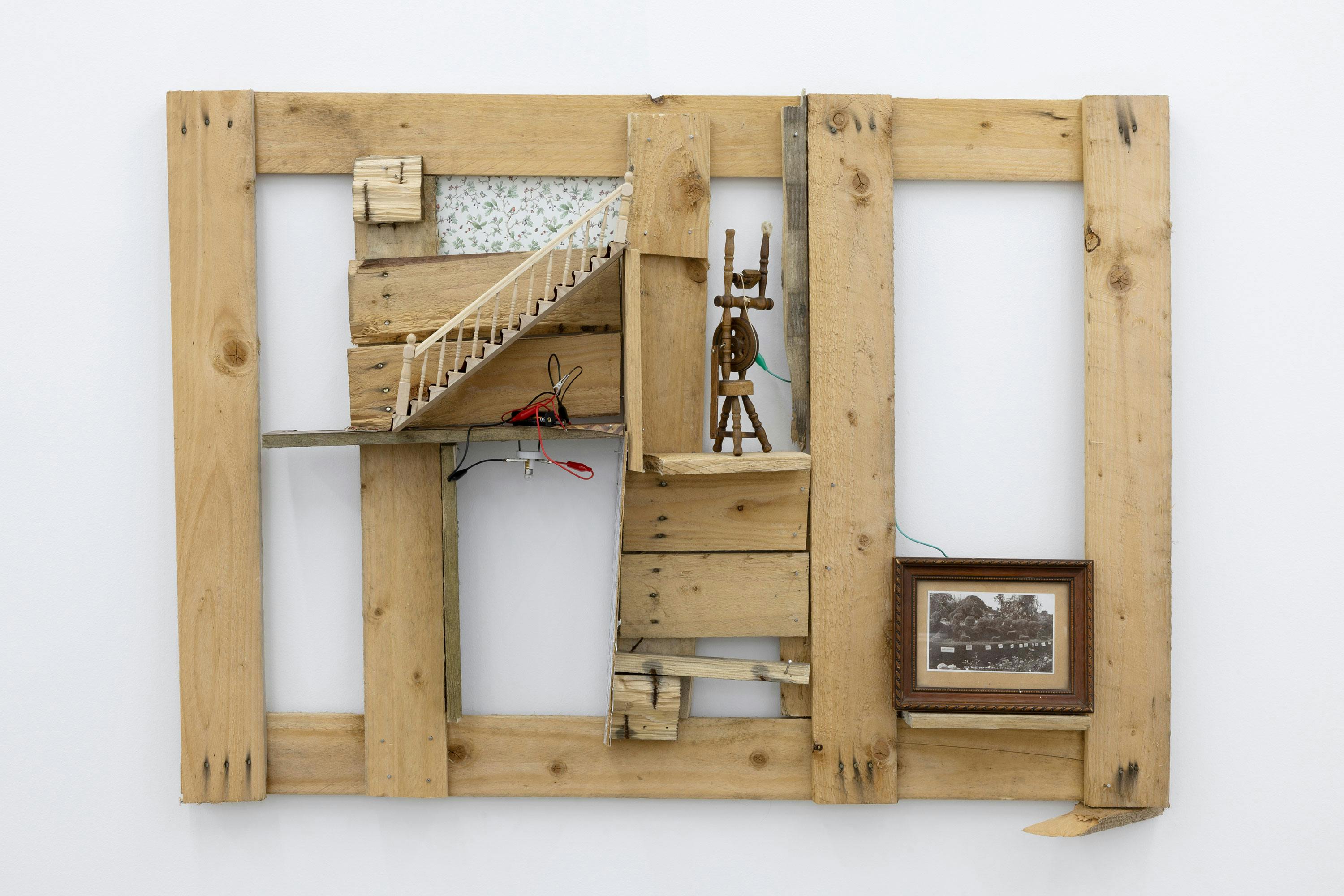 A wooden wall art piece with exposed panels, featuring a miniature staircase, a small wooden figure and a framed picture of a countryside scene. 