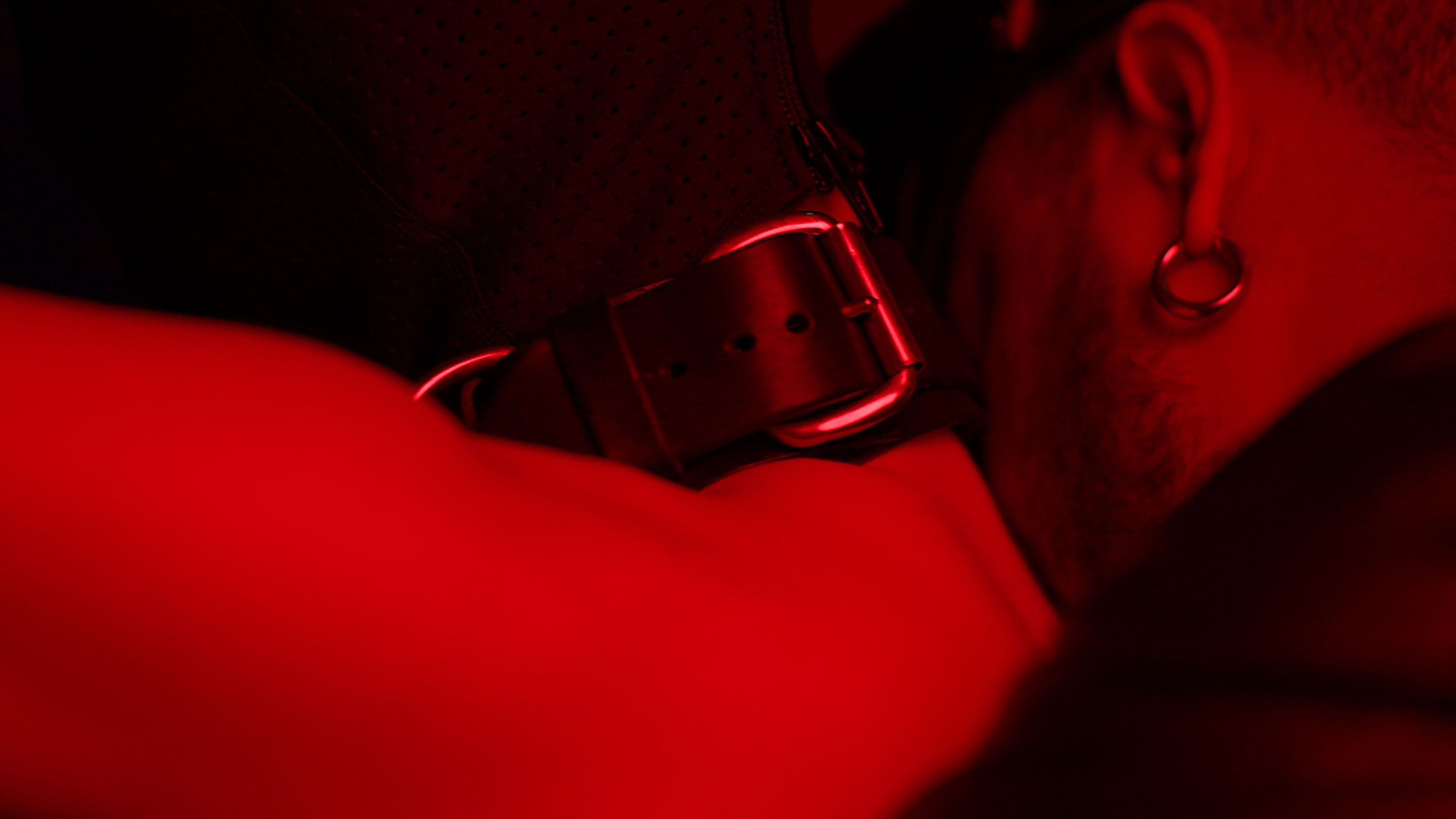 Close-up of a metal-buckled strap around a person's neck under dim red lighting. The person, partially visible, appears relaxed, with facial hair and an earring visible.