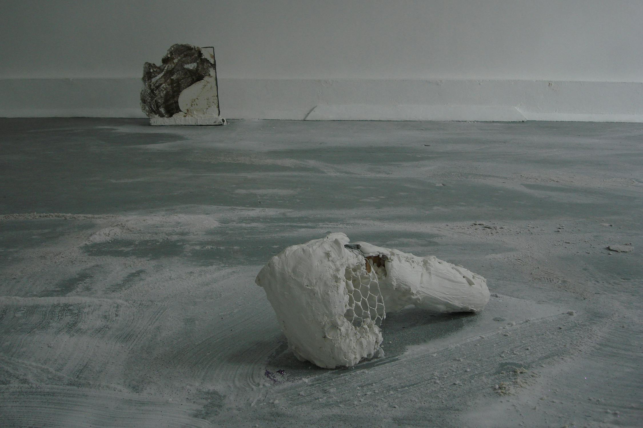 A minimalist art installation featuring two abstract sculptures. The foreground has a white, irregularly shaped piece with a wire, honeycomb texture. In the background, a rectangular piece leans against a white wall on a dusty grey floor.