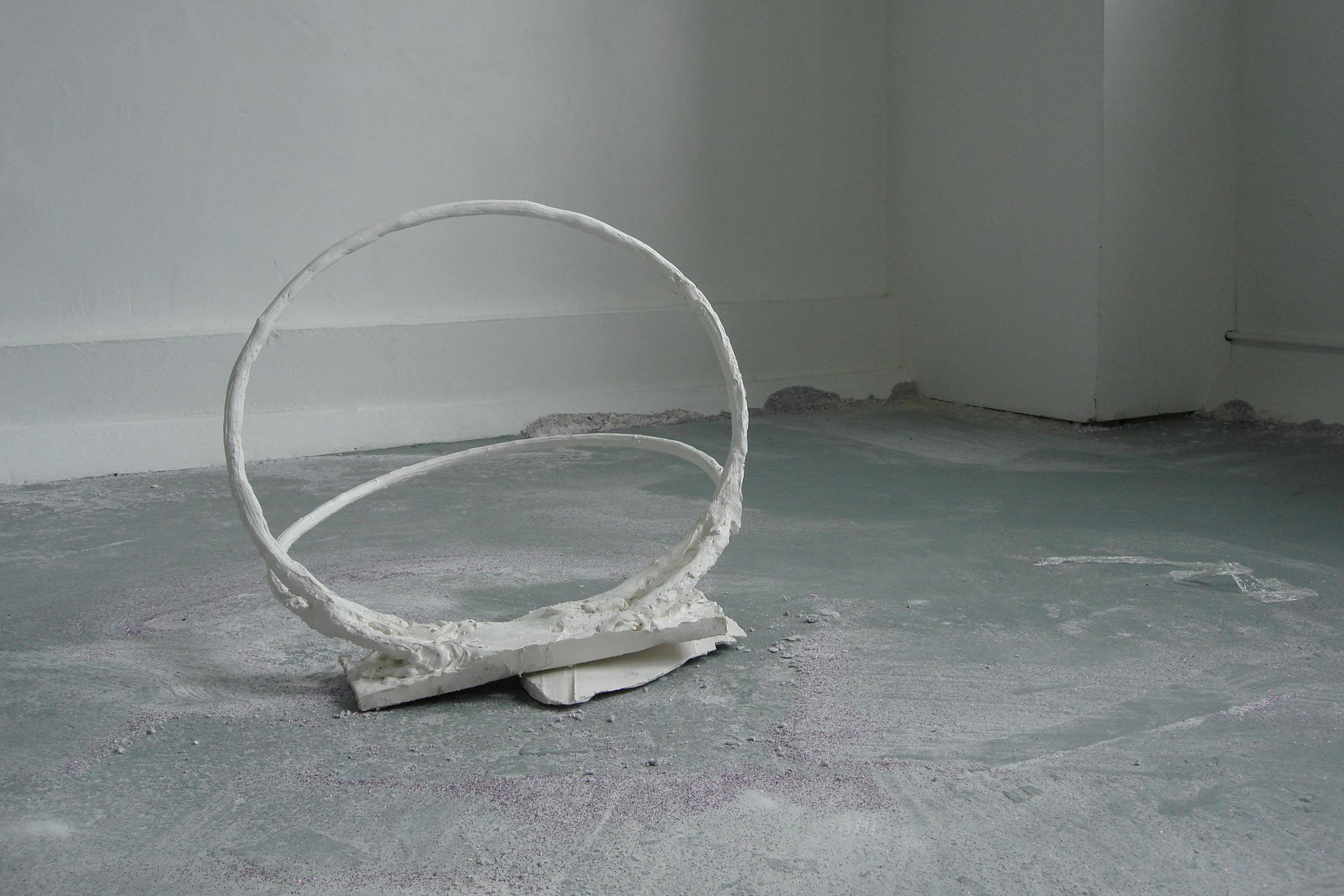 A white, round sculpture resembling a loop lies on a grey, textured floor in a minimalist room. The sculpture's base is irregular, with a small piece breaking the circle's continuity. The walls are plain white, and a dusting of purple glitter is scattered on the floor.
