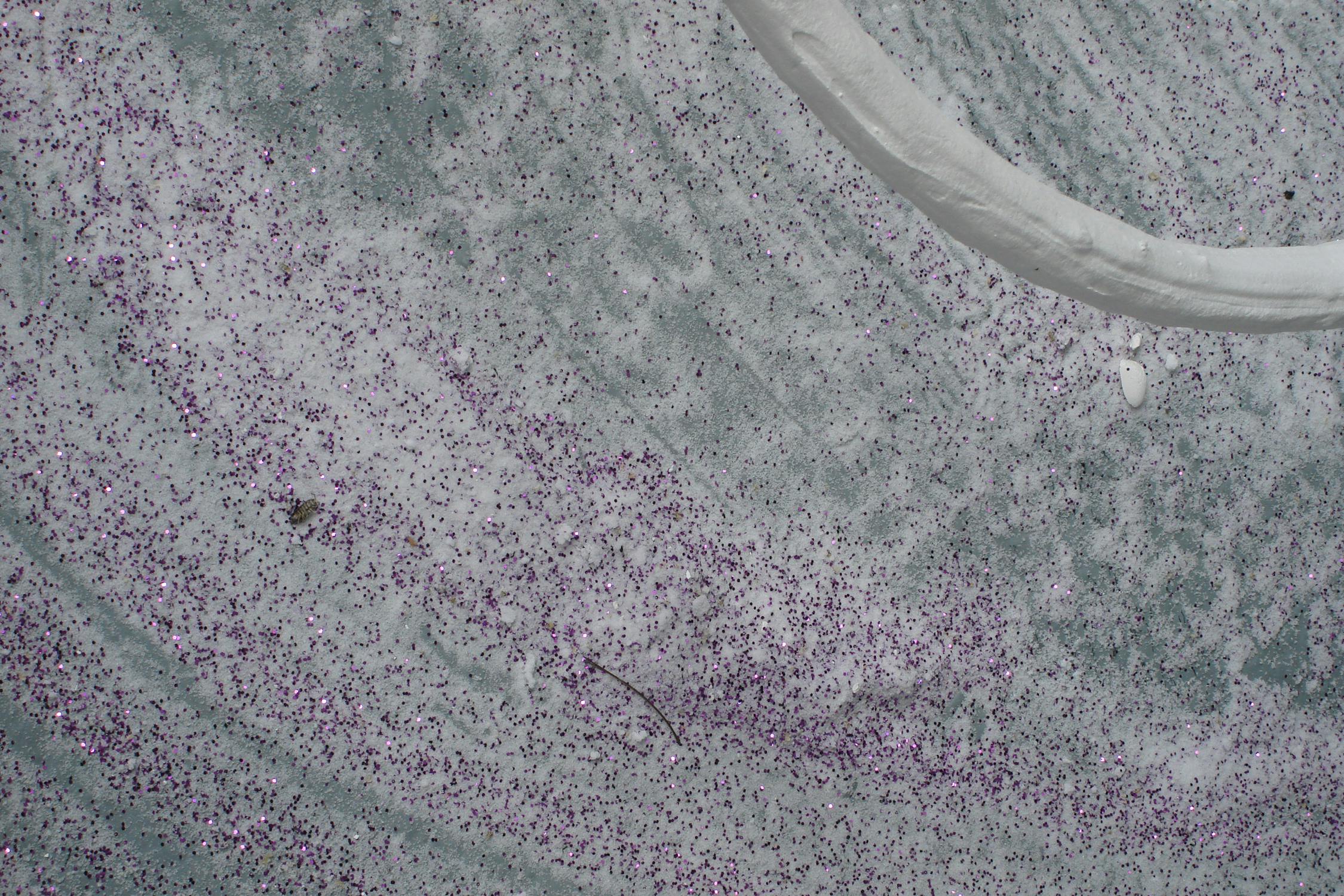 A close-up of a grey floor covered in a wash of white plaster with scattered purple glitter. A curved white looped sculpture crosses the upper right corner.