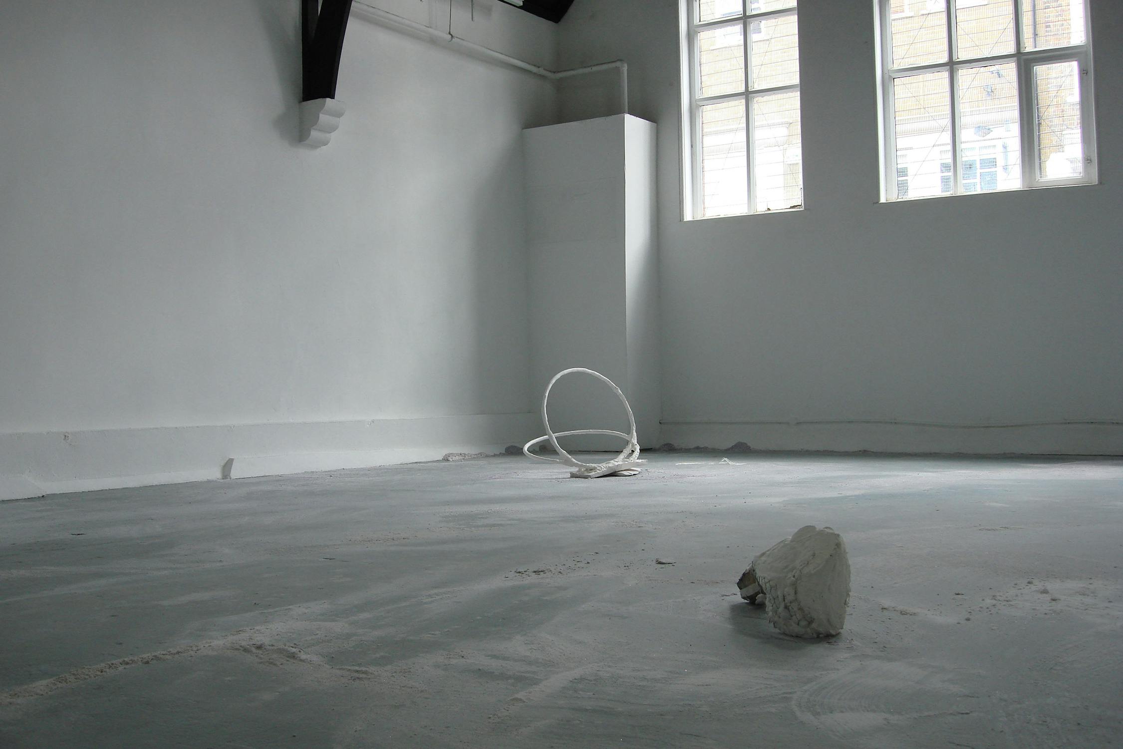 An empty, minimalistic room with white walls and a large window. There is a white coiled sculpture in the back corner and a small white object on the floor in the foreground. Natural light enters through the window.