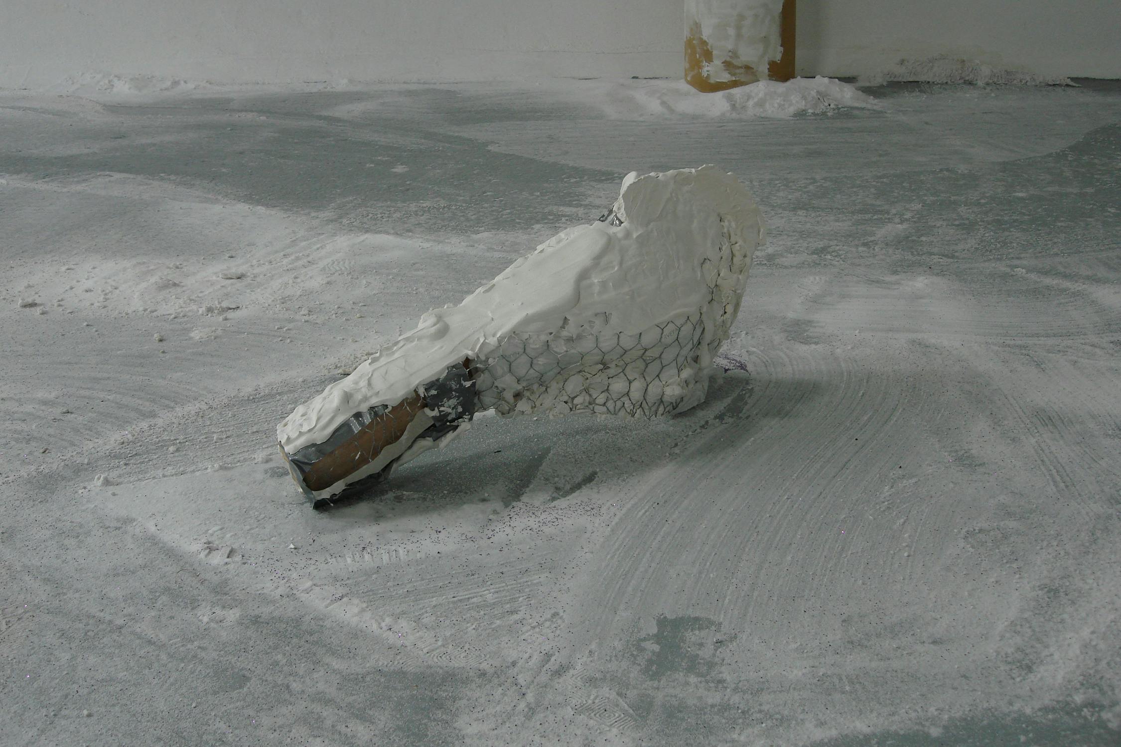 A sculpture resembling a crocodile's head, made from wire mesh and white material, perhaps plaster or modroc, rests on a textured, light grey floor. The surroundings are sparsely decorated, with white on the floor and a partially visible pole in the background.