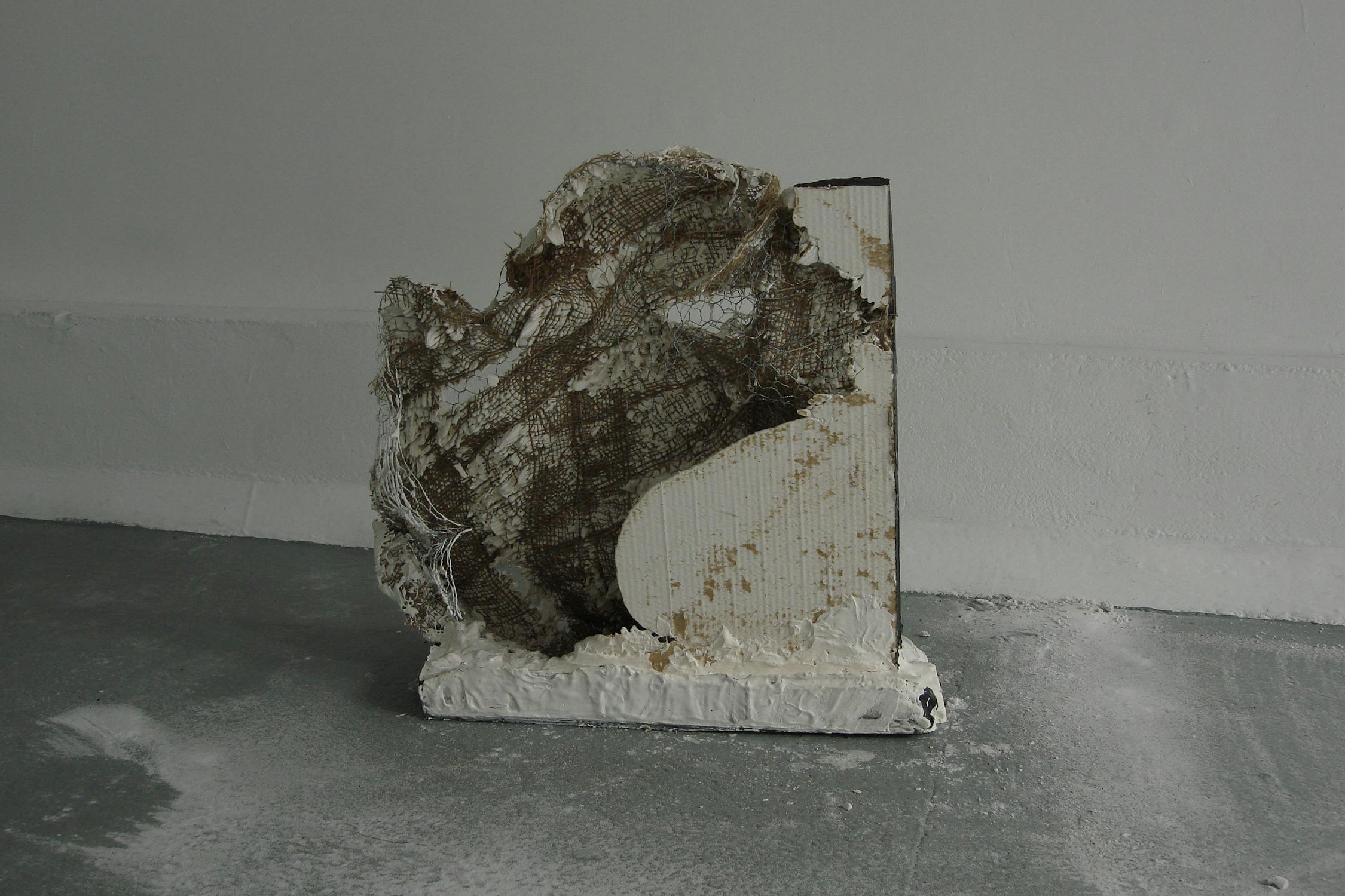 A textured sculpture with rough edges and a mix of white and brown surfaces is placed on a grey floor. The sculpture's irregular form seems to combine materials like plaster and mesh, creating an abstract and rugged appearance.