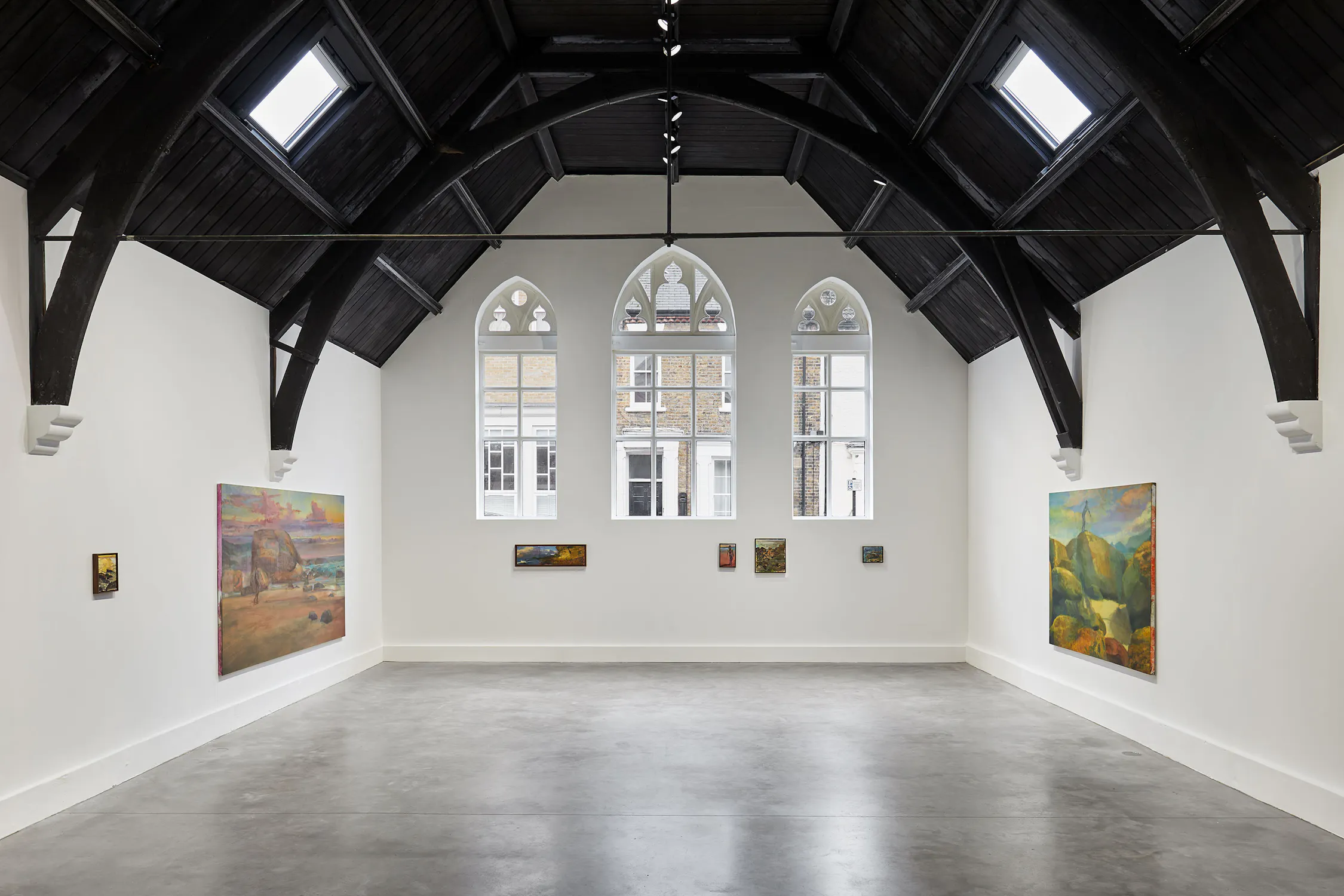 A spacious art gallery with high vaulted ceilings and arched windows. Several paintings by Jake Grewal are displayed on bright white walls. The gallery's minimalist interior features concrete flooring and exposed dark wooden beams.