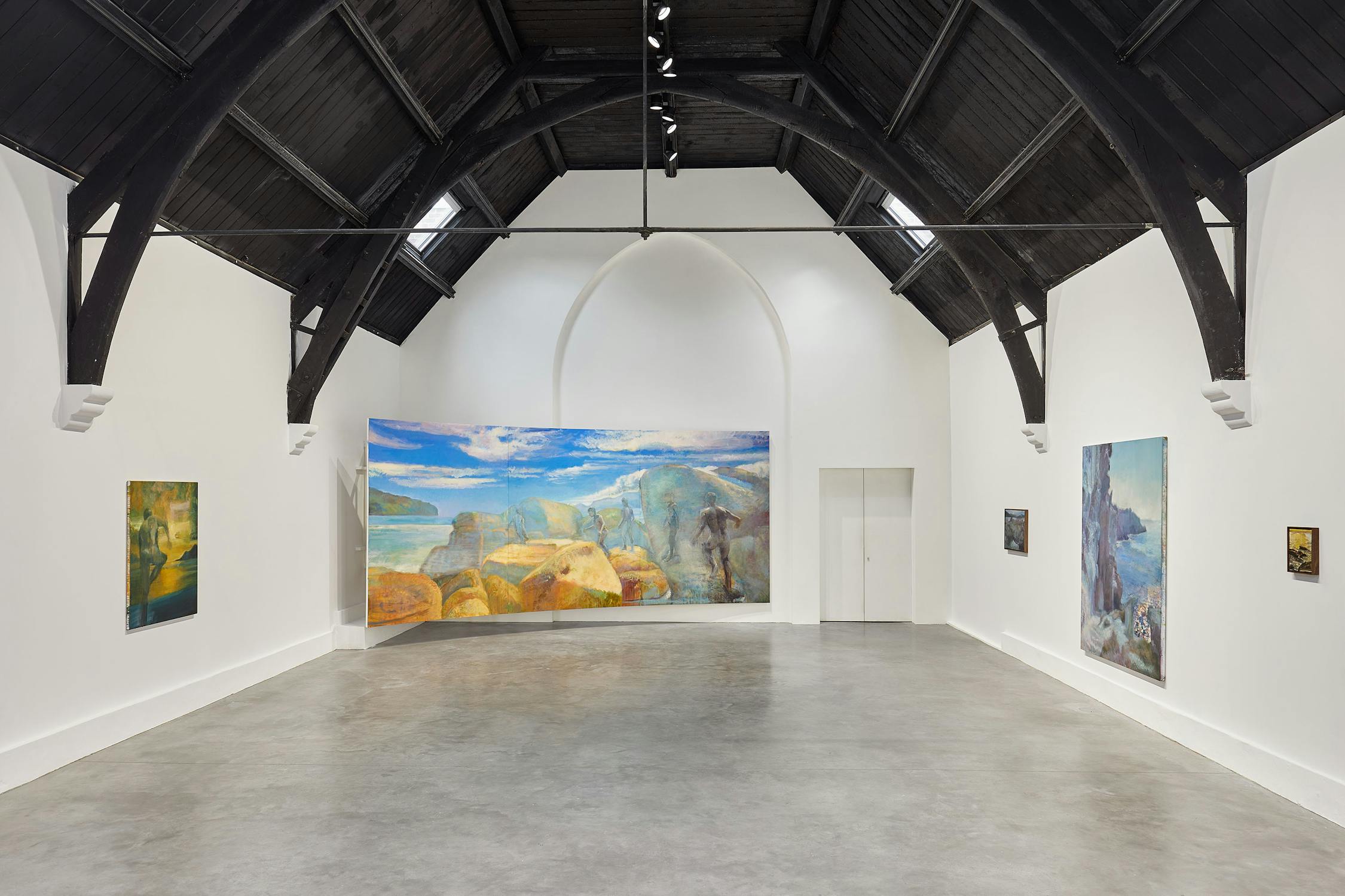 A spacious art gallery with a high, dark wooden ceiling and white walls. Several colourful paintings by Jake Grewal hang on the walls, with a large landscape painting prominently displayed at the centre. The floor is polished concrete.