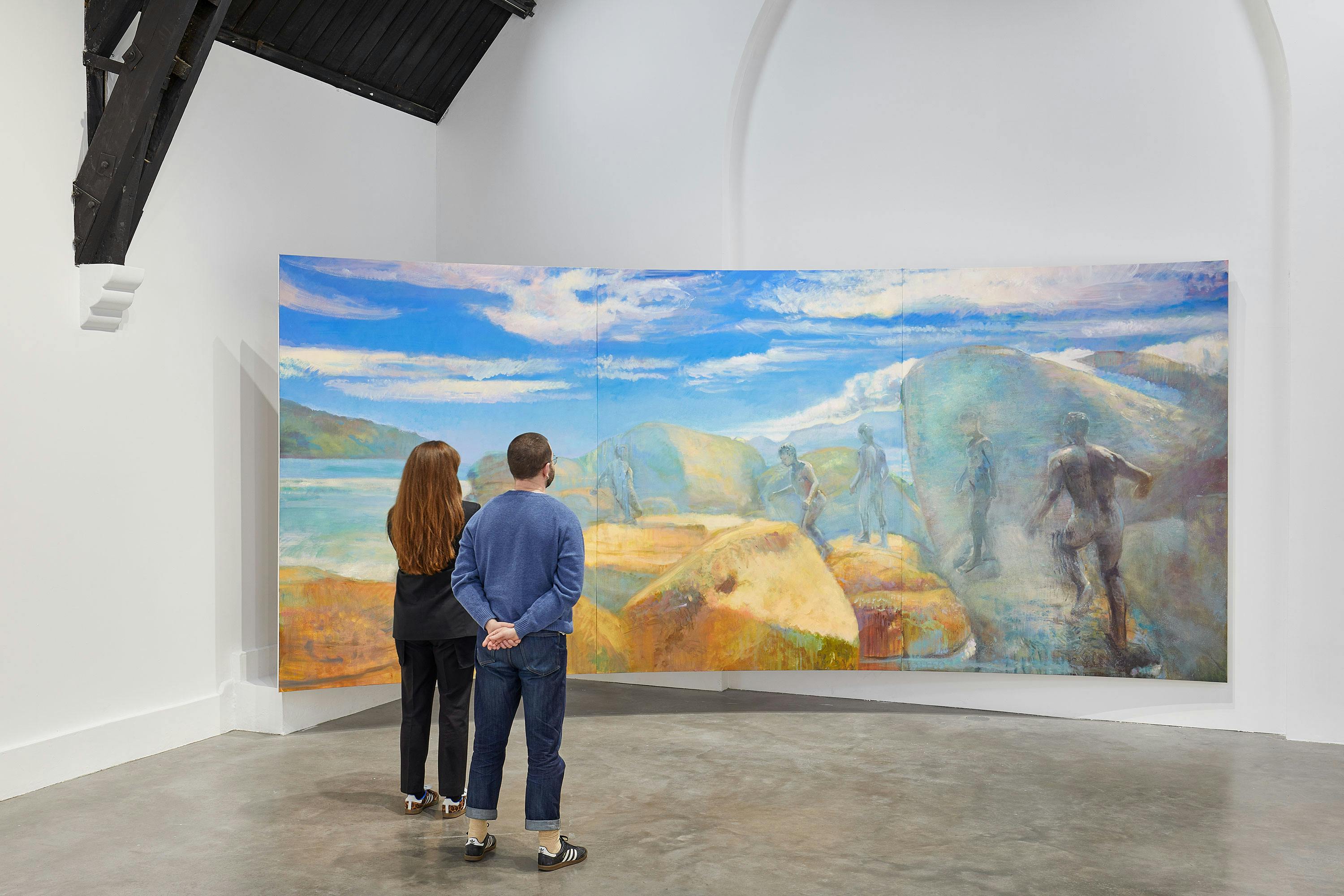 Two people stand in a gallery observing a large painting by Jake Grewal. The artwork depicts a landscape with rocks, water, and several figures under a blue sky with clouds. The gallery has white walls and a concrete floor.
