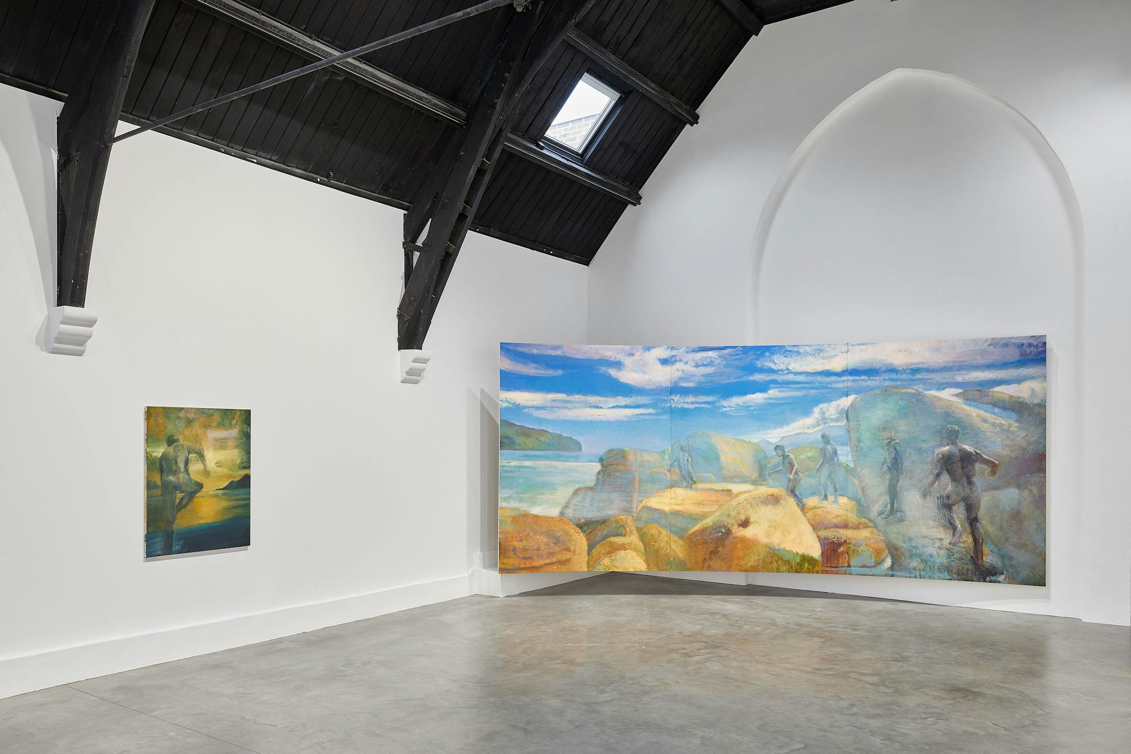 Art gallery with two paintings by Jake Grewal on white walls. The left painting depicts a sunset scene, while the right, larger painting shows figures on a rocky coastline under a bright blue sky. The room has a high, dark wooden ceiling with a skylight.