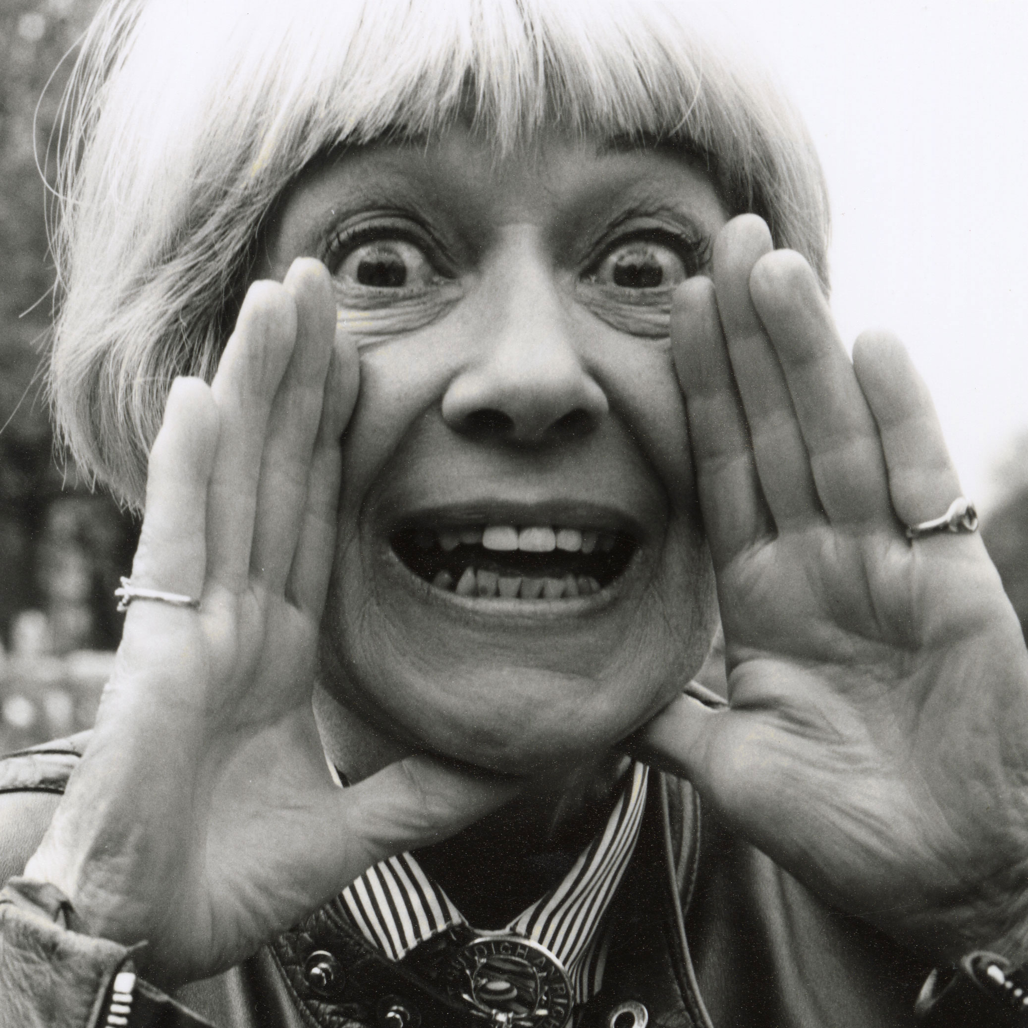 Portrait of Jackie Forster. She has a short blonde bob haircut holds their hands near their mouth, eyes wide open, and making a surprised or excited expression. She is wearing a leather jacket and have rings on their fingers. Black and white photo.