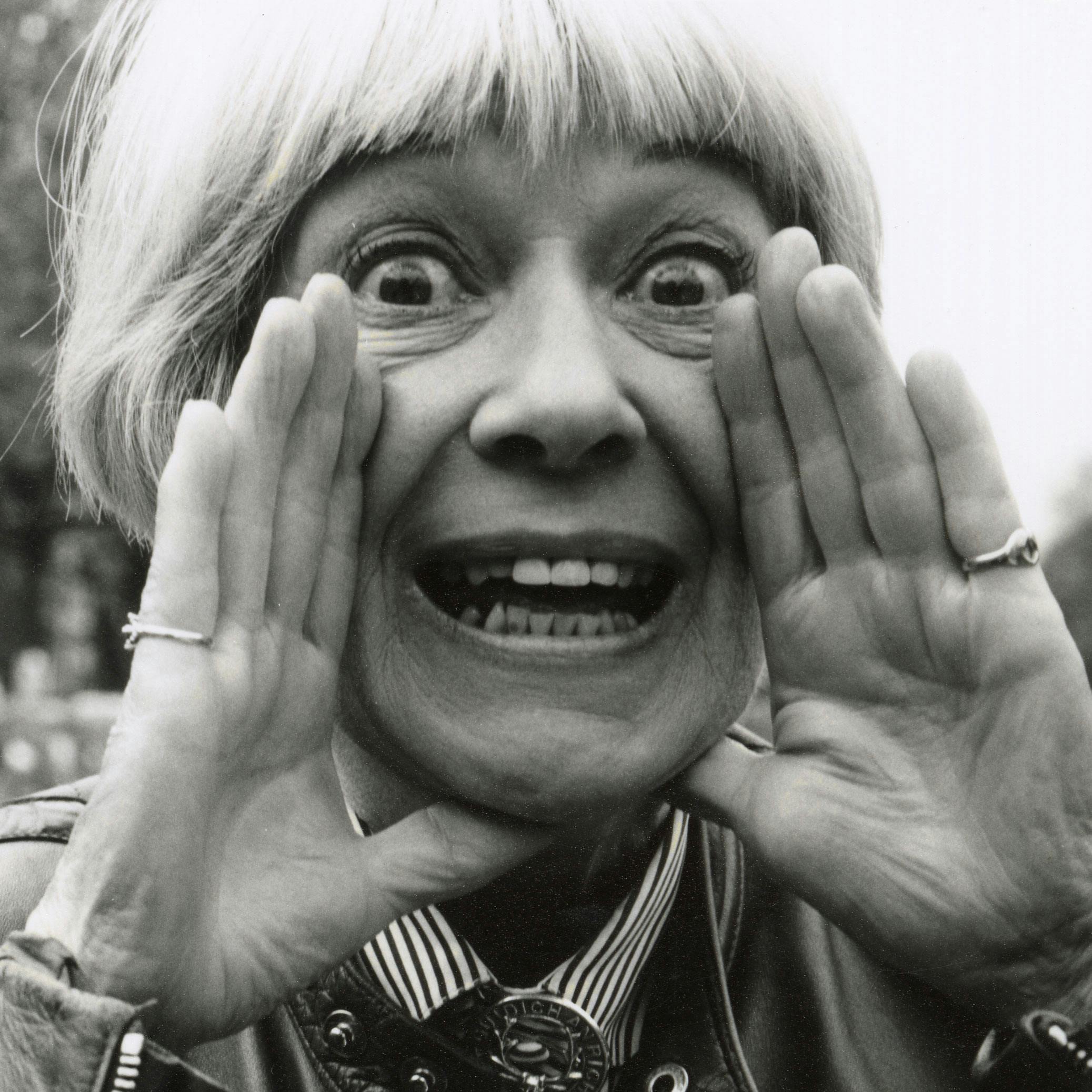 Portrait of Jackie Forster. She has a short blonde bob haircut holds their hands near their mouth, eyes wide open, and making a surprised or excited expression. She is wearing a leather jacket and have rings on their fingers. Black and white photo.