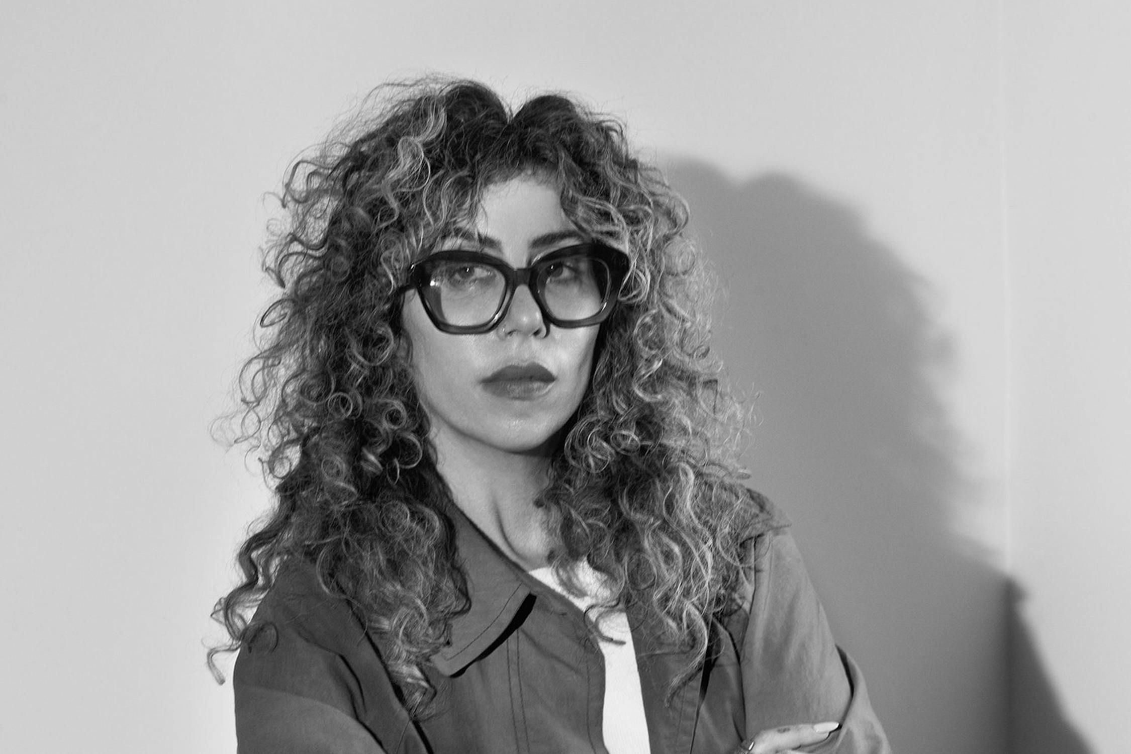 A black and white photo of Ariana Reines wearing large glasses and a jacket. They are looking out to the side of the camera with their arms crossed, against a plain wall.