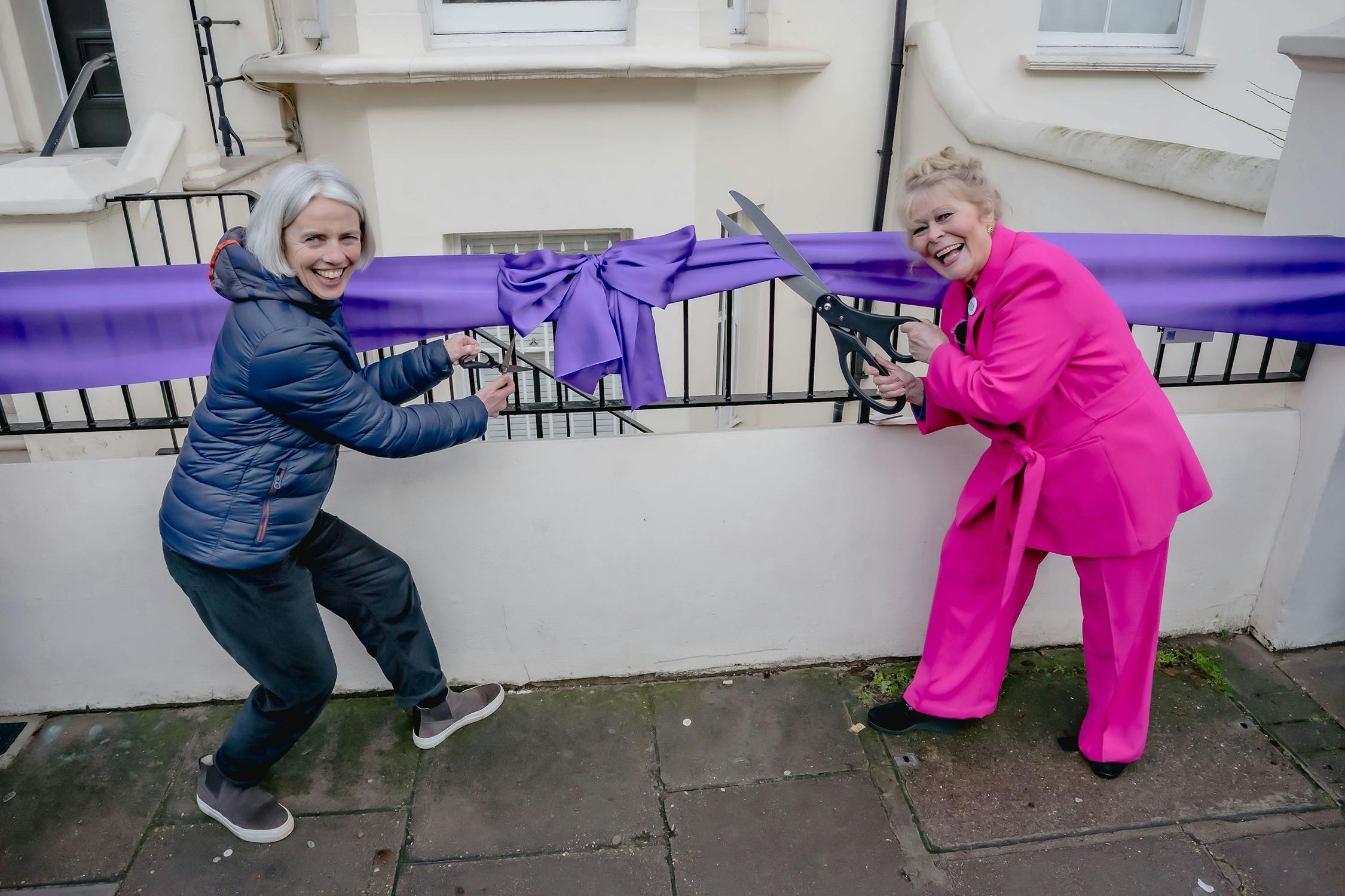 Two smiling people stand in front of a building, each holding a pair of large scissors as they prepare to cut a long purple ribbon tied in a bow along a fence. One wears a pink suit, the other a blue jacket and jeans.