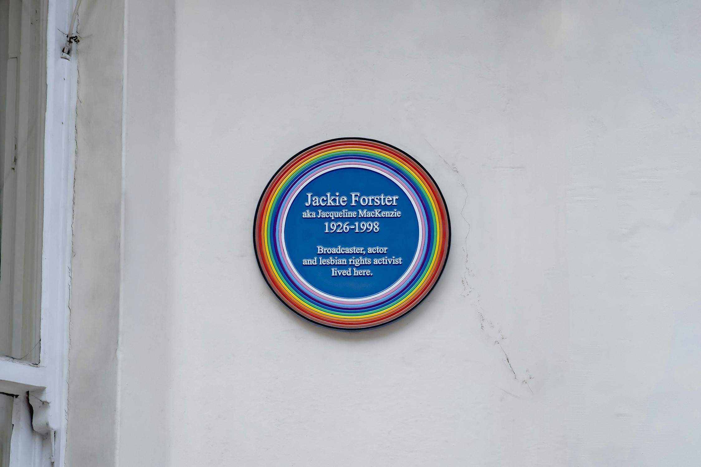 A blue plaque on a white wall, bordered by rainbow rings, reads: "Jackie Forster aka Jacqueline Mackenzie 1926-1998 Broadcaster, actor and lesbian rights activist lived here.