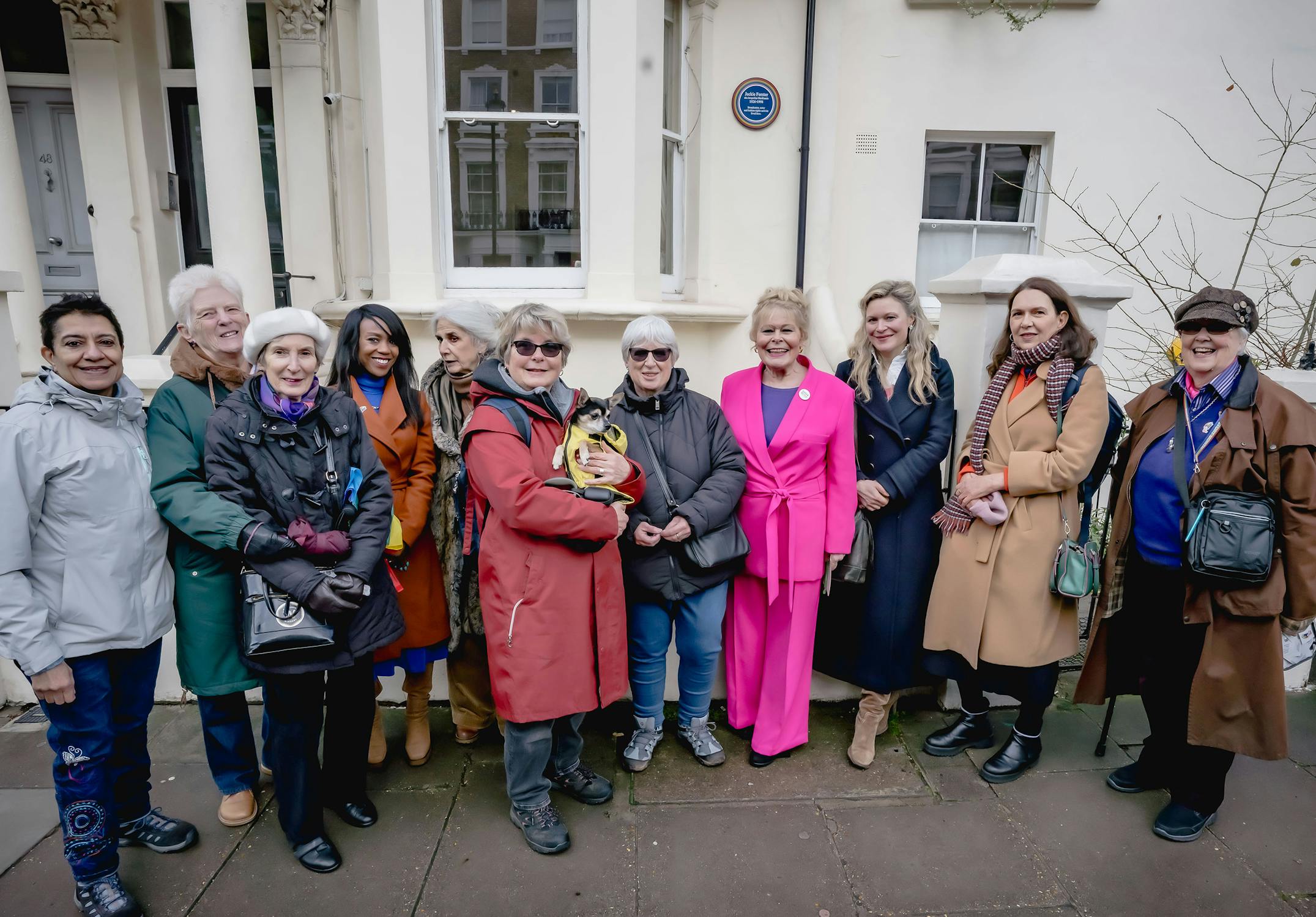 A group of people standing closely together on a pavement, smiling for the photo in front of a building with white columns and a blue plaque. One person holds a small dog. The clothing style varies, with some wearing coats.