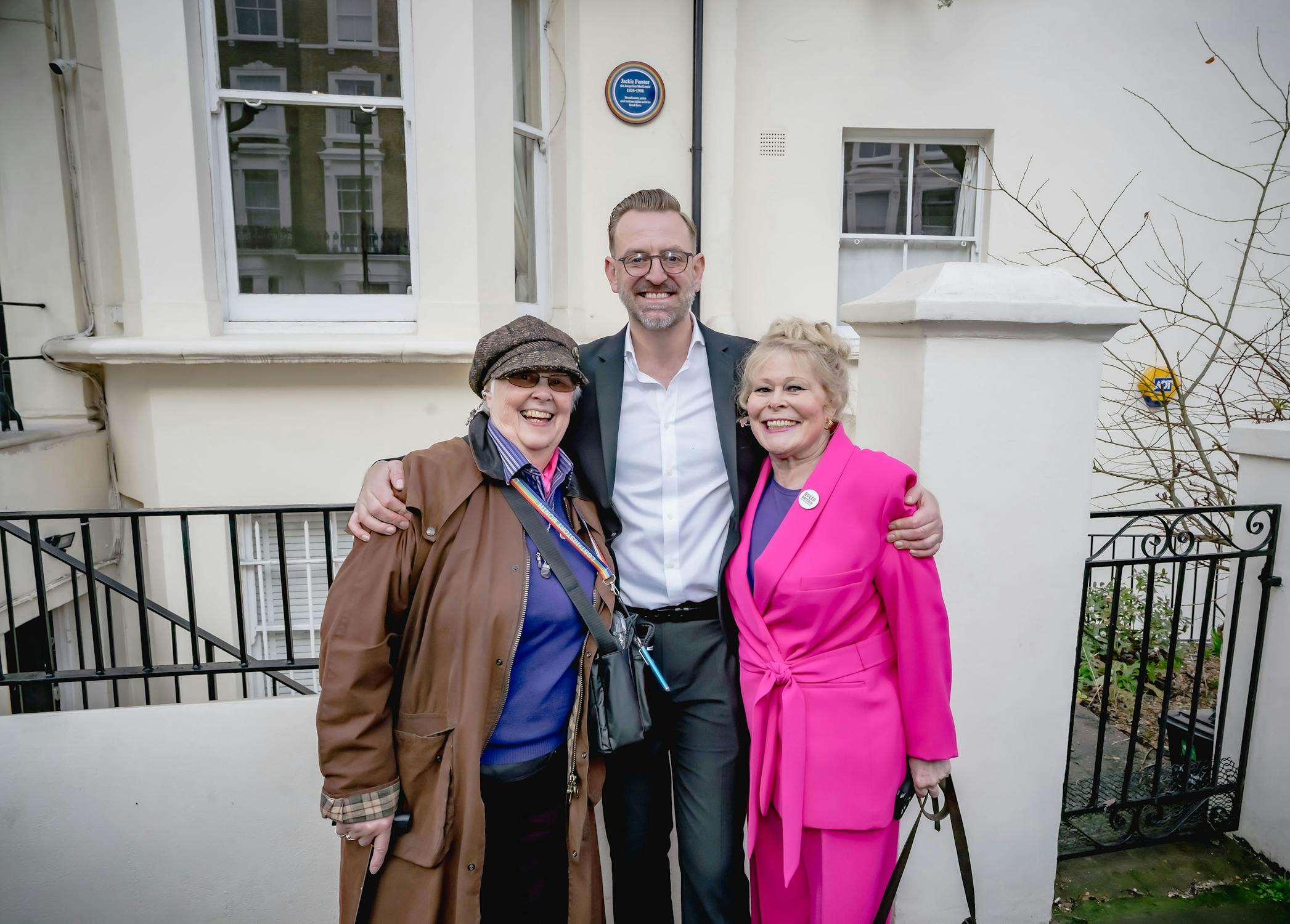 Three people stand smiling in front of a white building. The person on the left wears a brown coat and hat, the person in the middle wears a suit, and the person on the right wears a bright pink outfit. They are posing with their arms around each other.