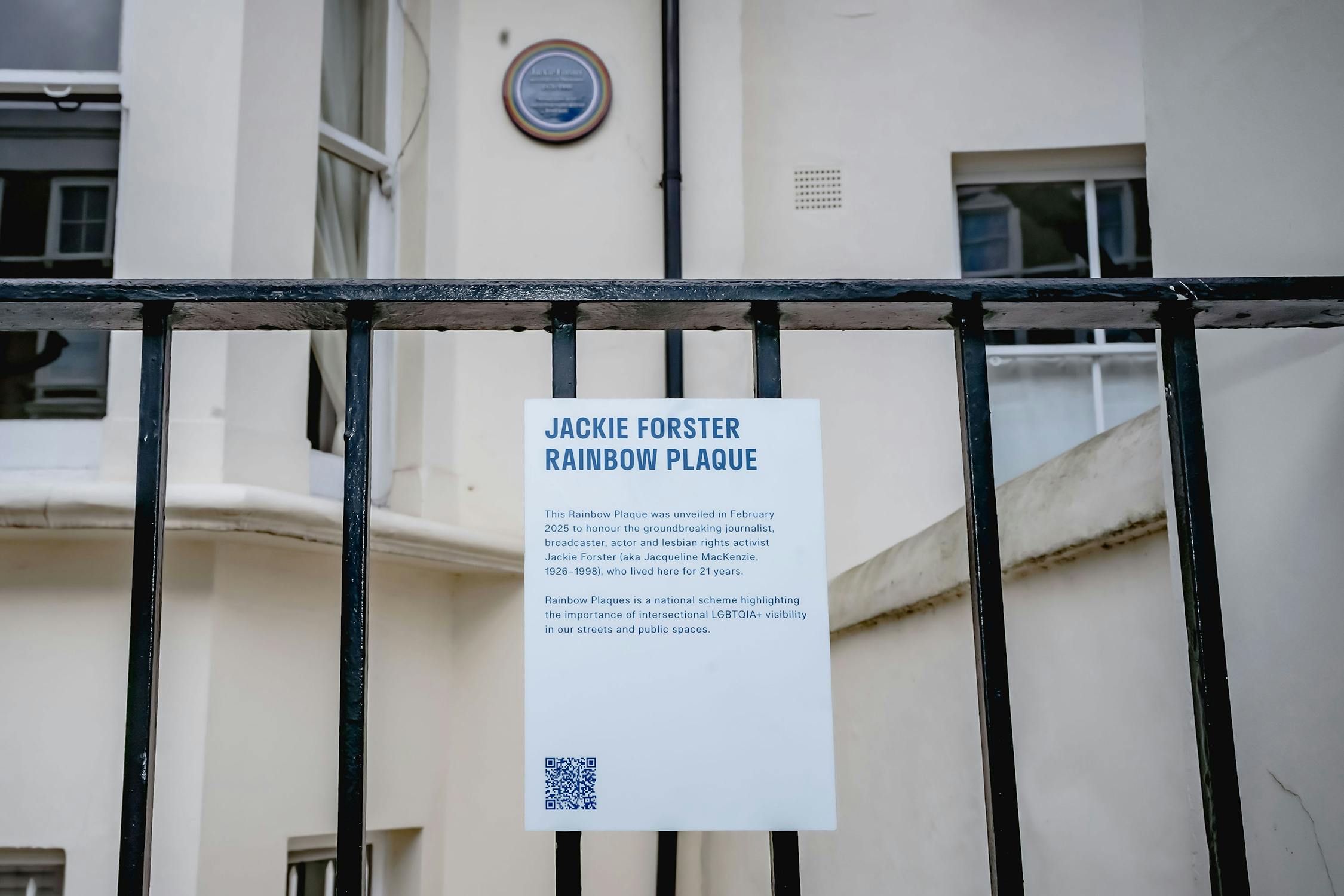 A white sign behind a black metal fence reads “Jackie Forster Rainbow Plaque.” It explains the plaque honours her contributions to LGBTQ+ visibility. A blue plaque is mounted on the building wall above. A QR code is present on the sign.
