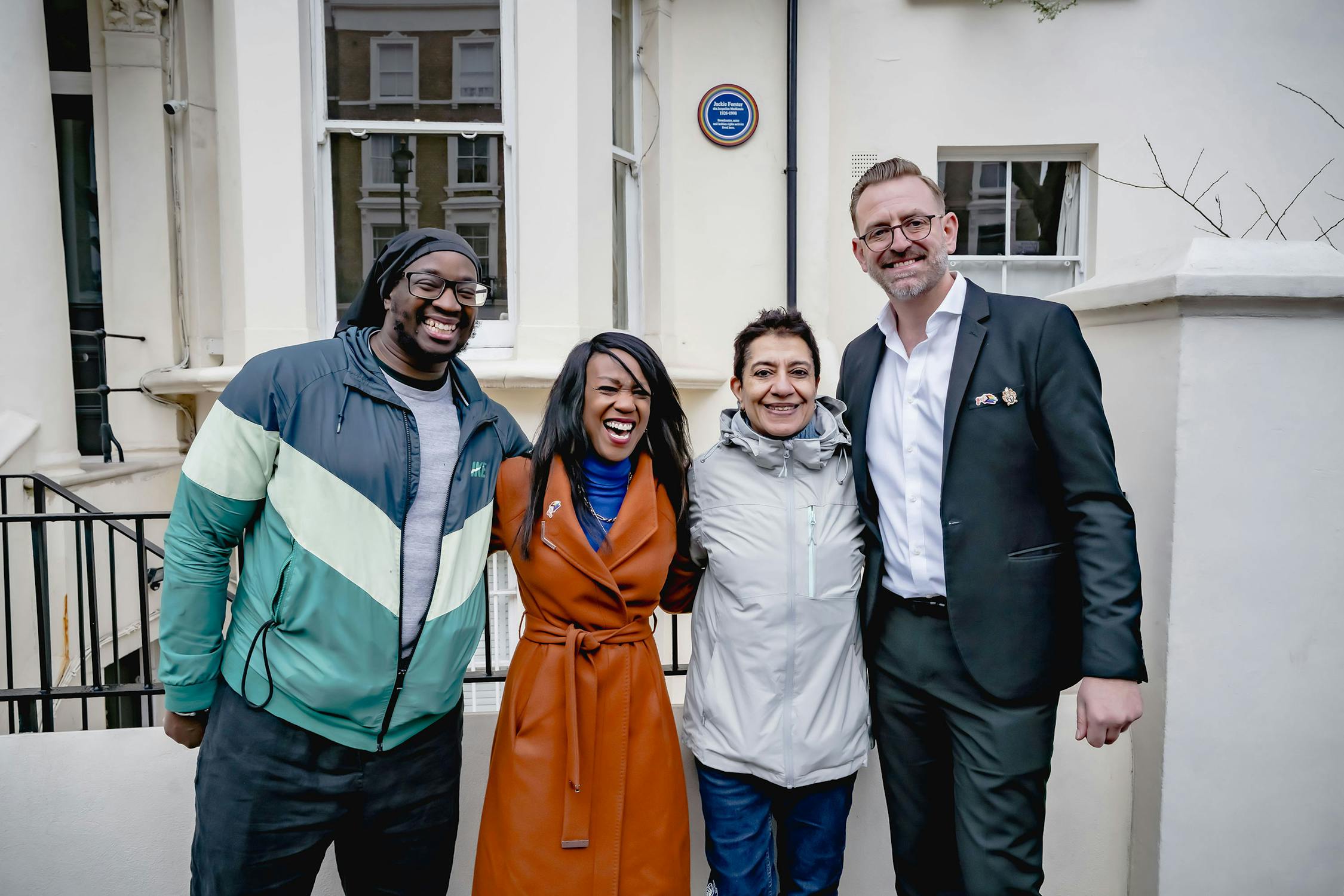 Four people stand together outside a building's entrance. They are smiling and wearing jackets. A blue plaque is visible on the wall behind them. The building is cream-coloured with large windows and white trim.
