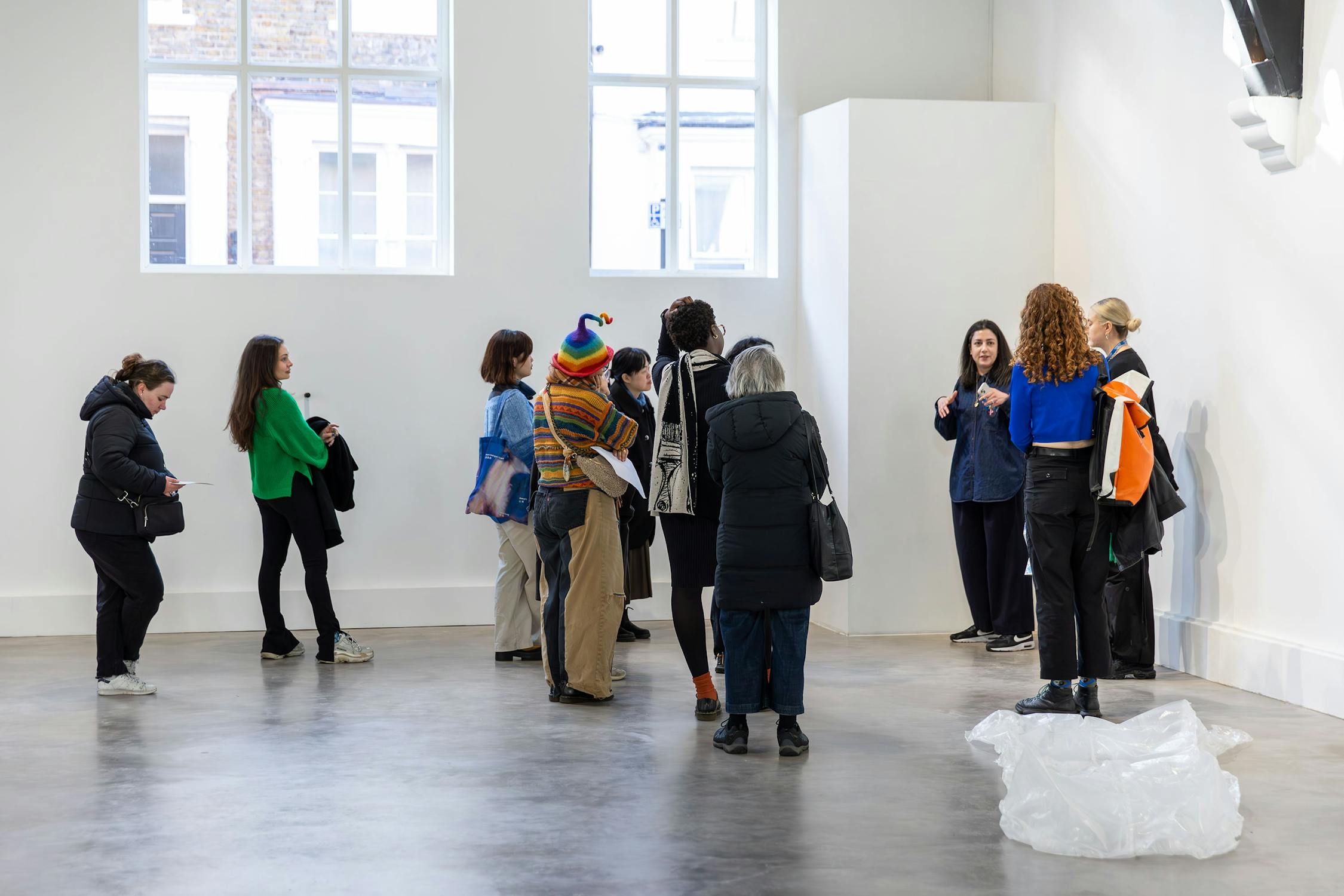 A group of people in a modern, minimalist art gallery. They are gathered around a piece of art near a white wall, listening to a person speaking. Light floods in from large windows, and some artwork is visible in the background.