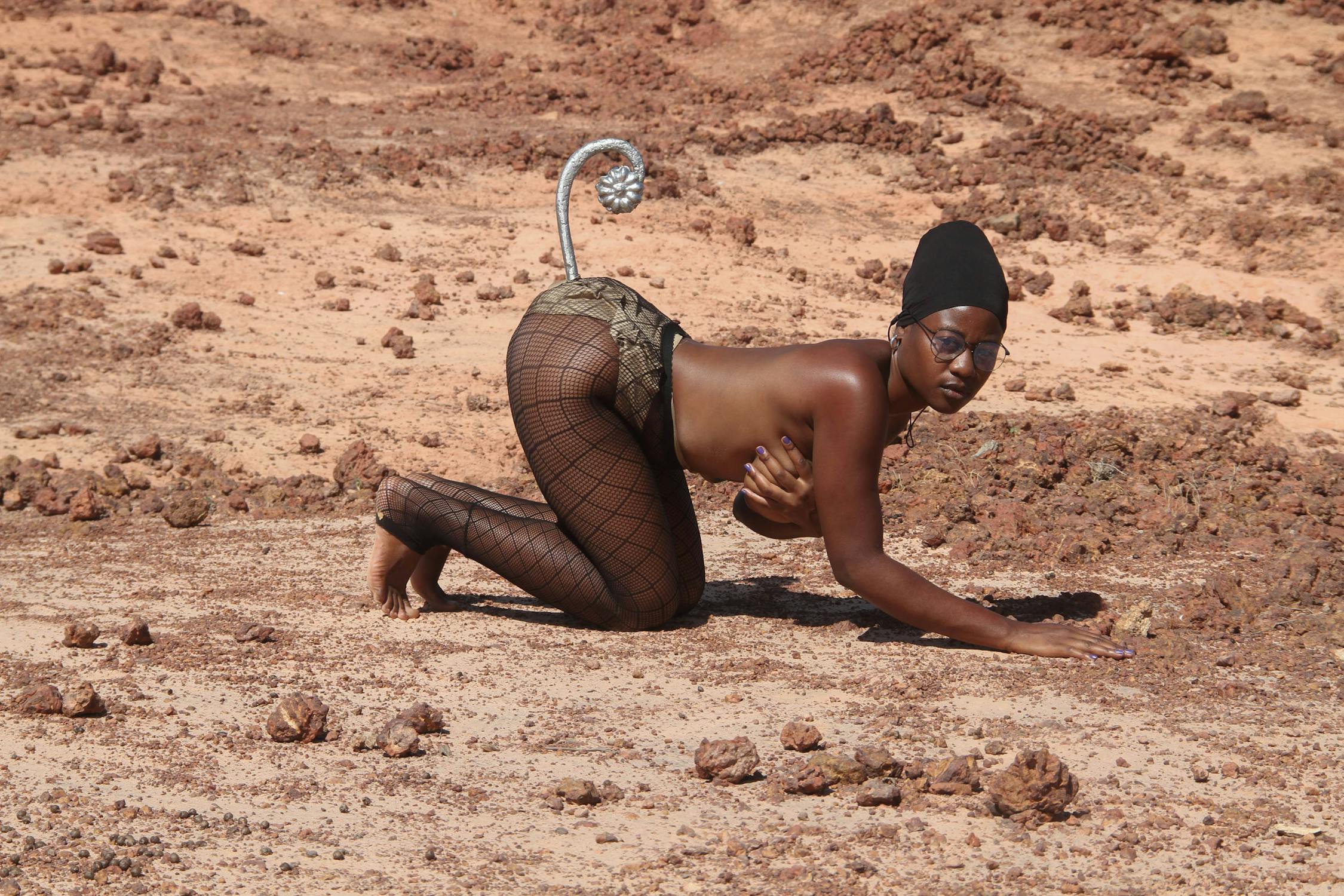 A person poses on all fours in a desert-like landscape, wearing netted leggings, snakeskin shorts, and sunglasses. A metal, tail-like accessory is attached to the waist. The setting is sandy with scattered rocks.