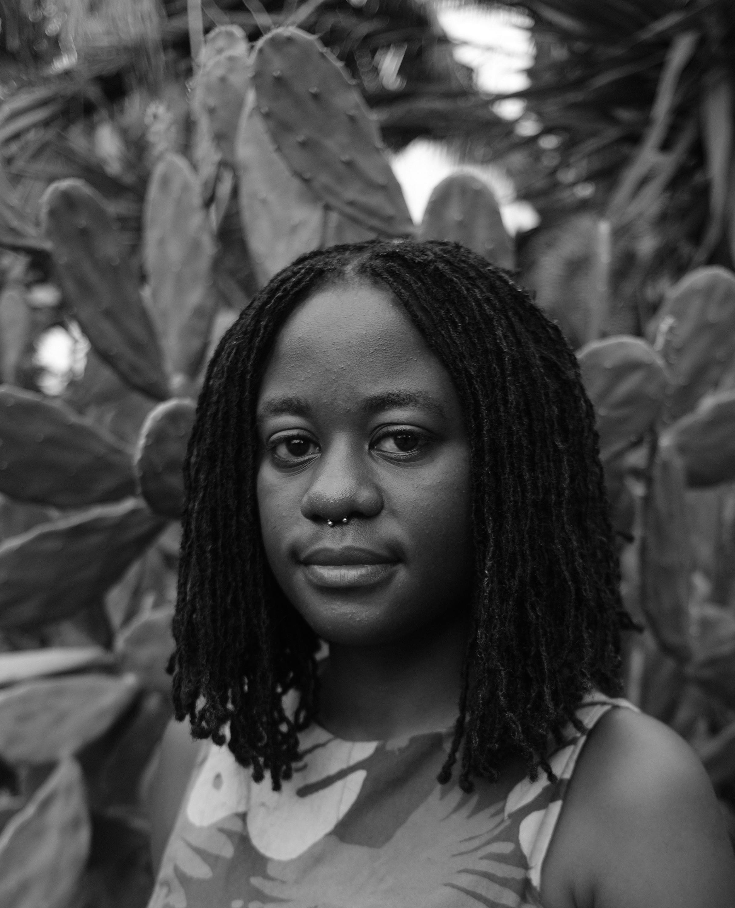 Photographic portrait of artist Godelive Kasangati Kabena. She has shoulder-length braided hair and a nose ring. Kabena stands in front of large cactus plants, wearing a sleeveless patterned top. The image is in black and white.