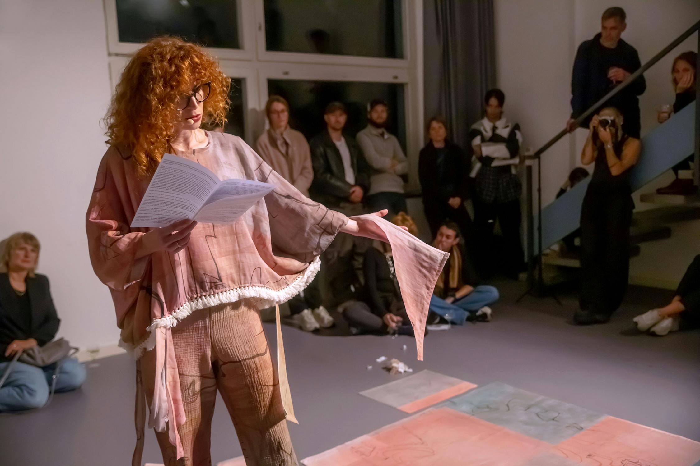 A person with curly red hair is reading a paper to a group of seated and standing audience members in an indoor venue. They're wearing a flowing, textured garment. The audience is attentively watching, and artworks are displayed on the floor.
