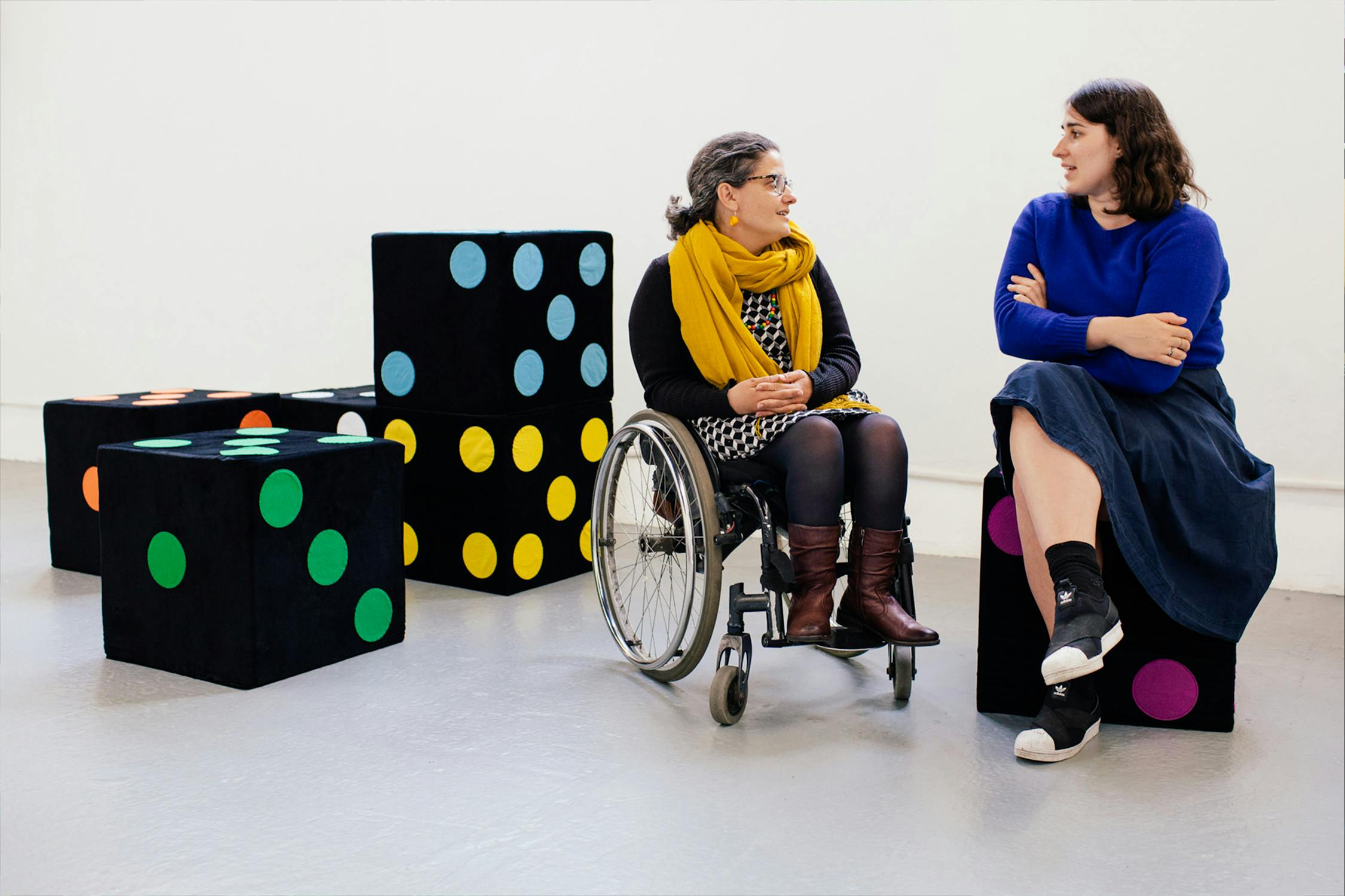 Two people are sitting and talking, with one in a wheelchair. They are surrounded by large black cubes featuring colourful dots. The person in the wheelchair wears a scarf and boots, while the other wears a blue sweater and skirt.