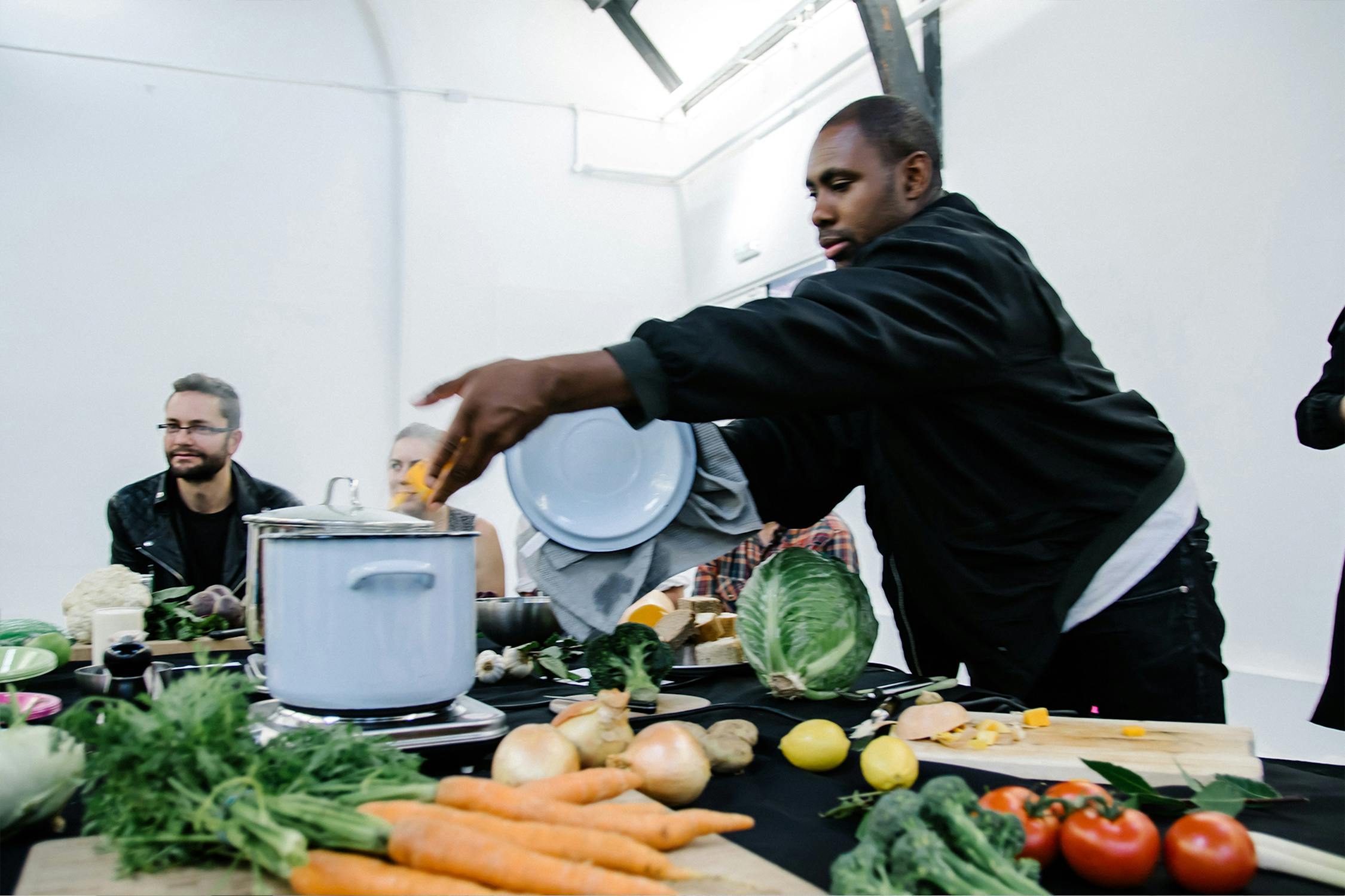 A man in a kitchen setting places a lid on a pot surrounded by various vegetables, including carrots, tomatoes, and cabbage. Three other people sit at the table observing the activity. The room is bright with a modern, minimalistic design.