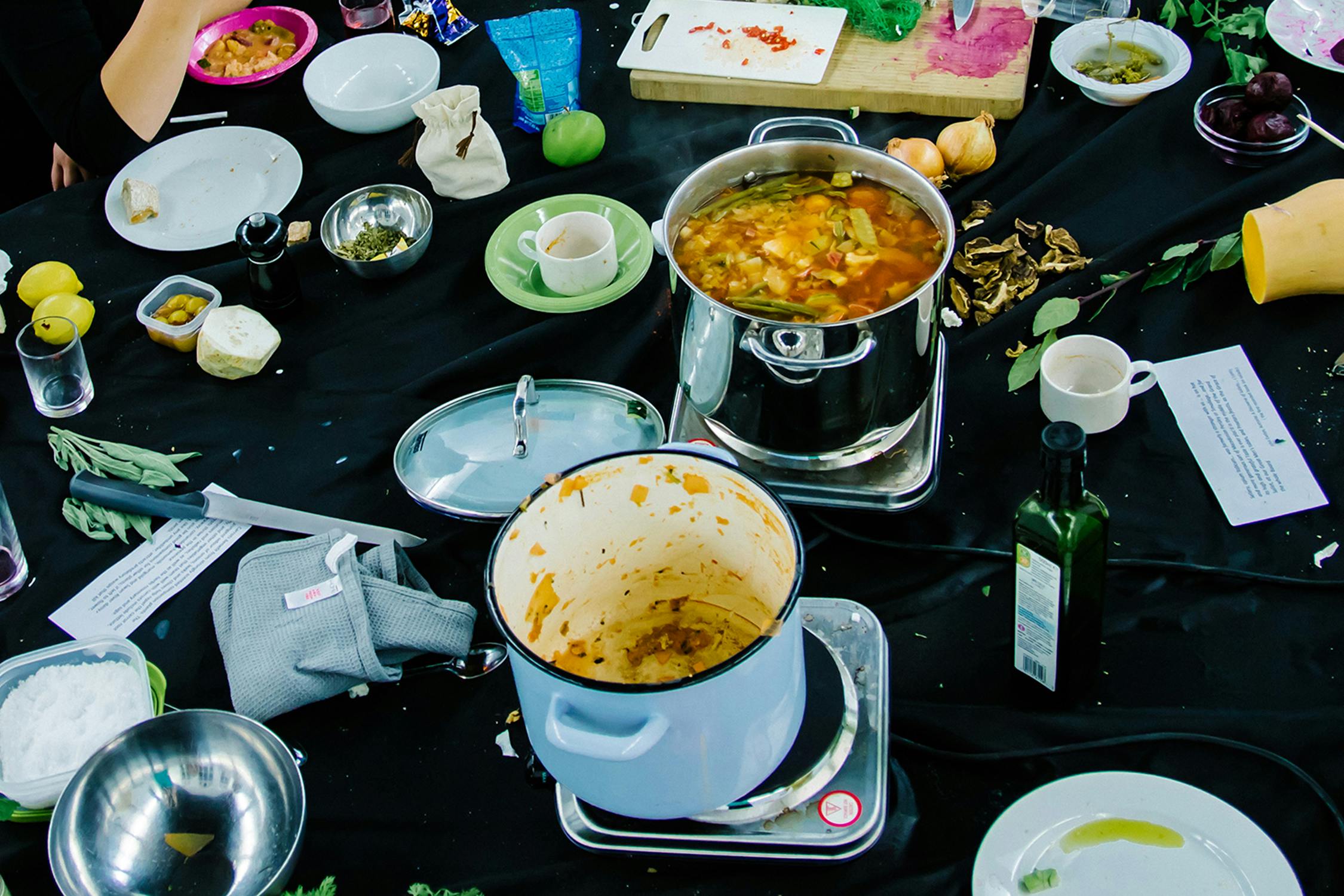 A table covered with a black tablecloth is filled with cooking items. There are two pots, one full of soup and one empty, various herbs, spices, vegetables, and used plates and cups scattered around.