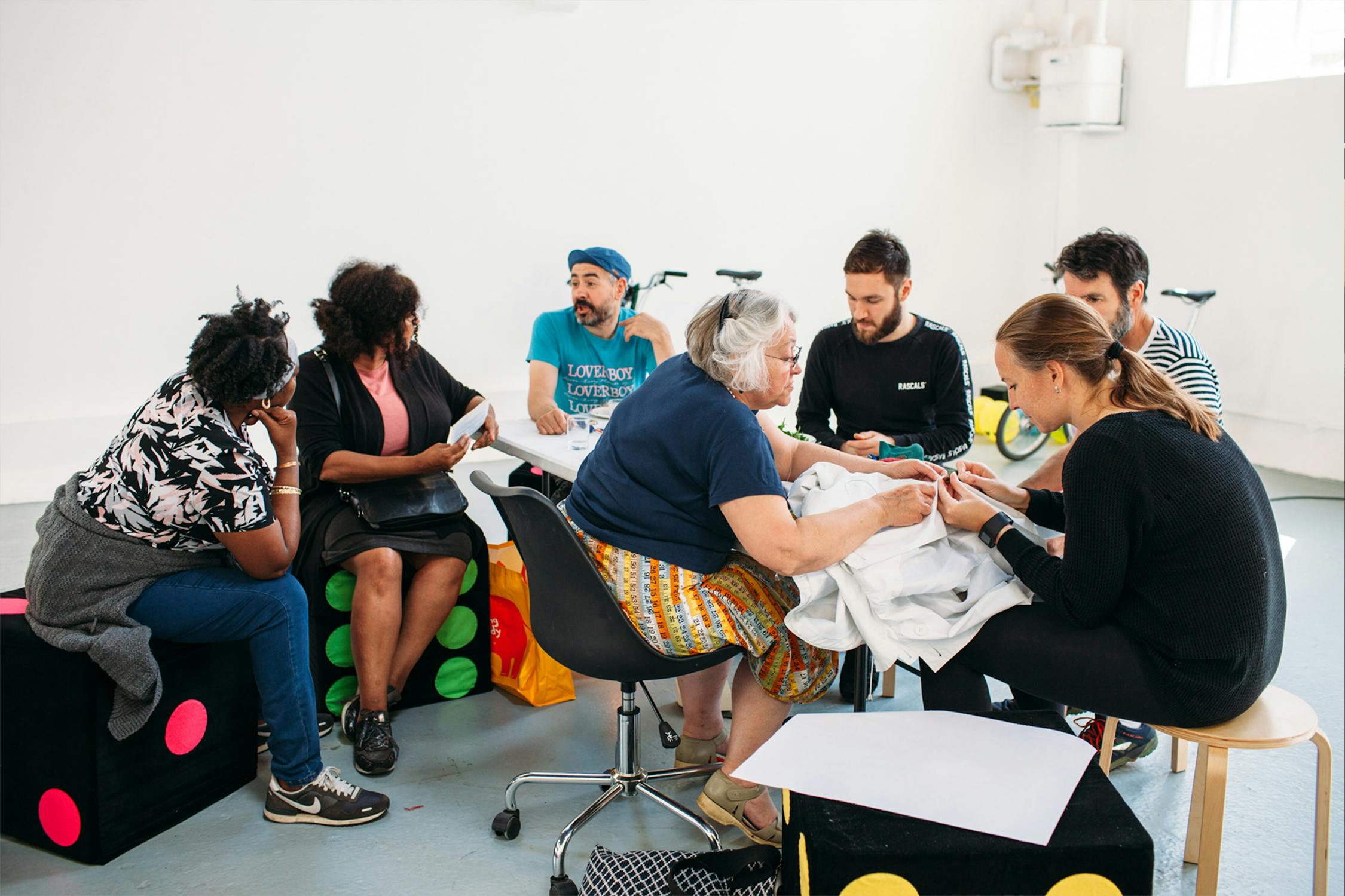 A diverse group of people are sitting in a circle, engaging in a group activity. They are seated on colourful stools and chairs, focusing on an object on the table in the middle. The room is bright and has bicycles in the background.
