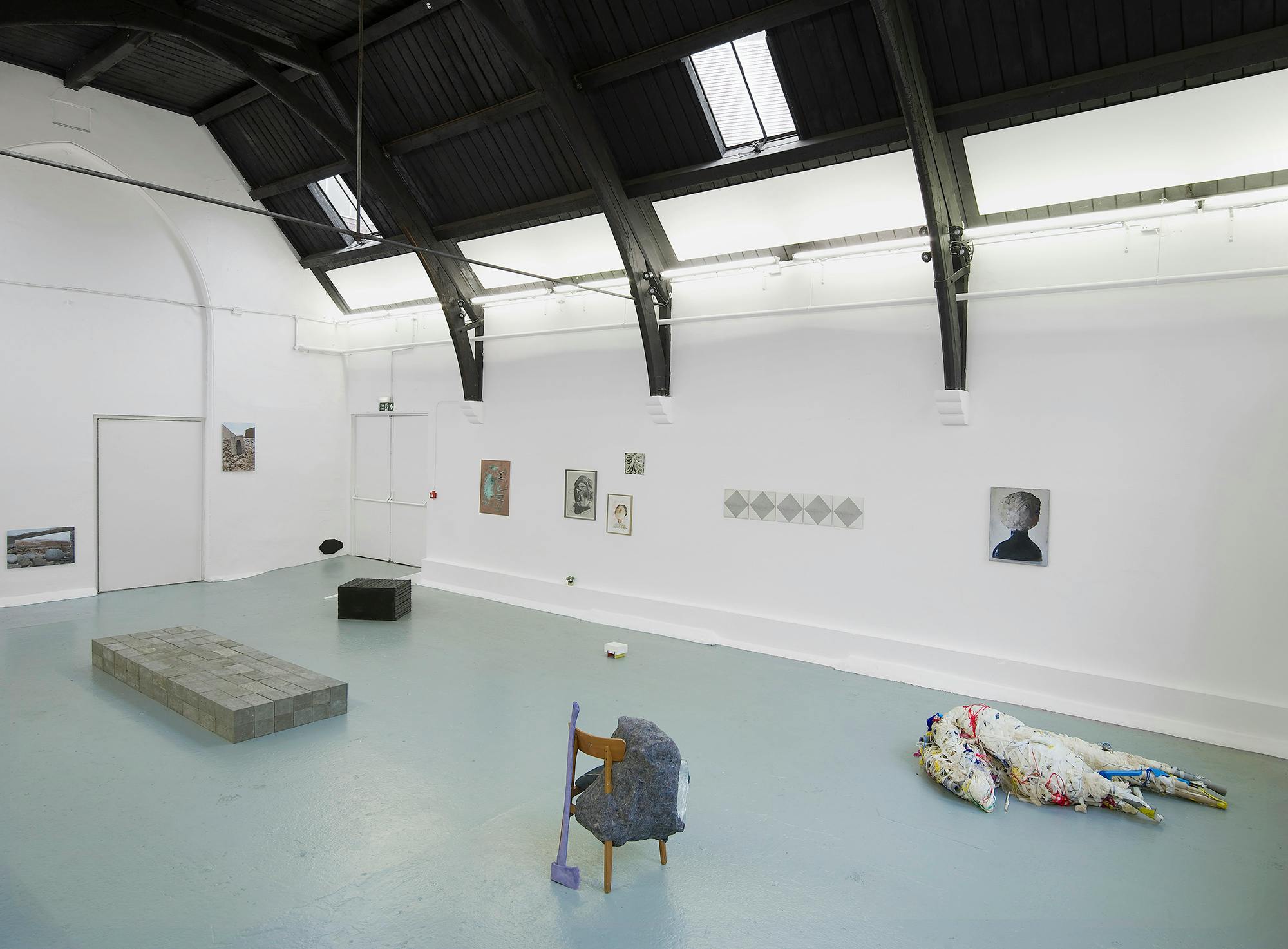 A contemporary art gallery with white walls and black ceiling beams displays various artworks, including paintings on the walls, a brick platform, a small black block, a chair with fabric, and a fabric-wrapped figure on the floor.