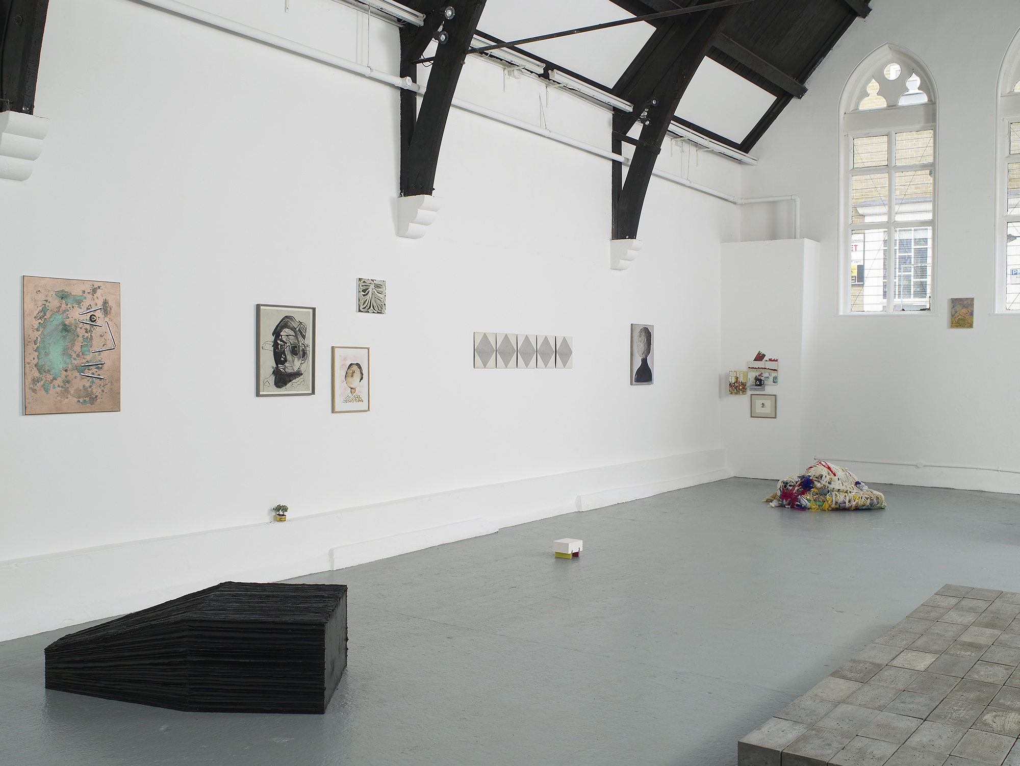 A contemporary art gallery with white walls and high, vaulted ceilings displays sculptures, paintings, and photos. Artworks are placed on the floor, plinths, and walls, in a minimalist, spacious arrangement.