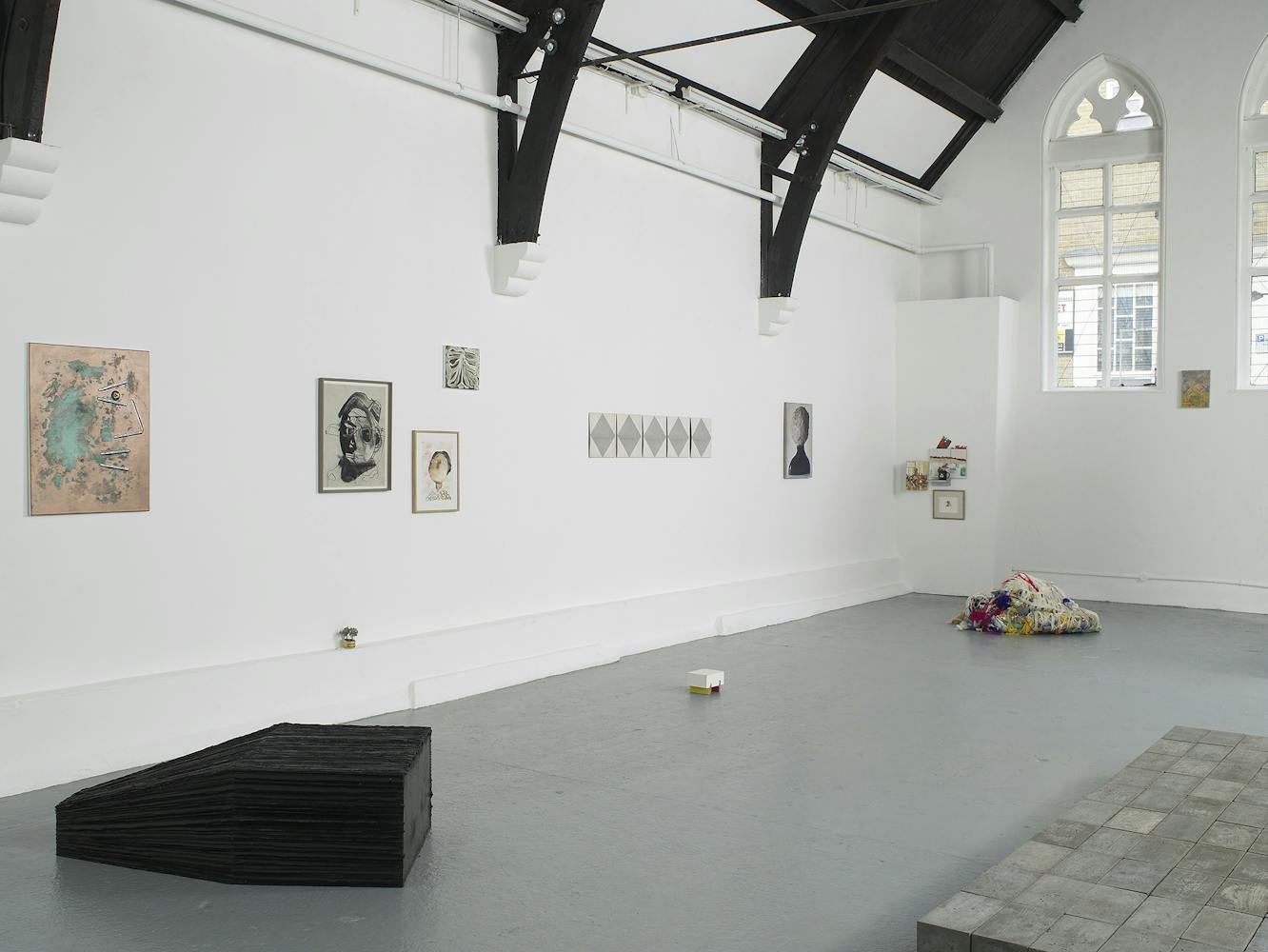 A contemporary art gallery with white walls and high, vaulted ceilings displays sculptures, paintings, and photos. Artworks are placed on the floor, plinths, and walls, in a minimalist, spacious arrangement.