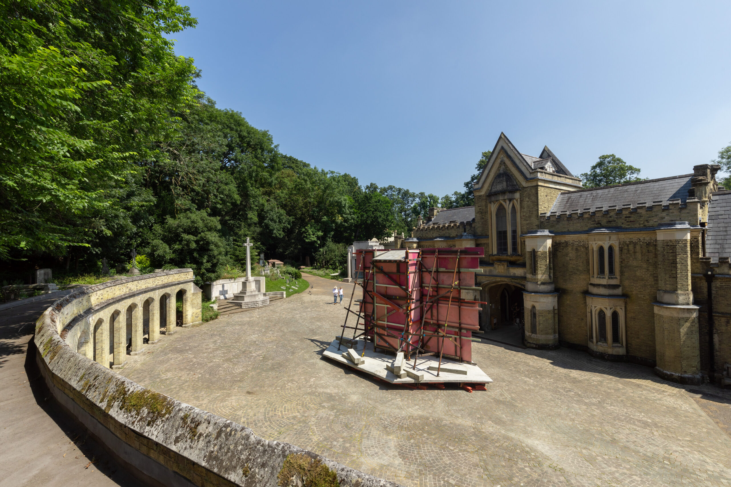 A paved courtyard with a stone colonnade curves left, leading to a historic, gothic-style building on the right. In the centre, a large red and white scaffolding structure stands. Trees and a tall white monument are in the background under a clear blue sky.