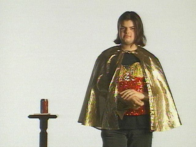 A person with long hair stands against a white background, wearing a shiny gold cape over a black and red sequined shirt. They are next to a small stand with a soda can on it.